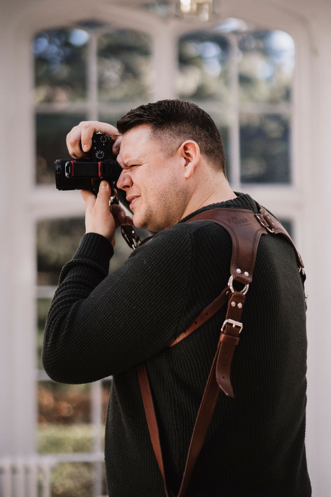 A photographer in a black sweater and leather harness strap takes photos during a wedding.