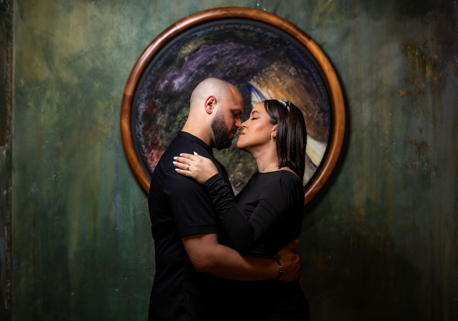 A couple shares an intimate moment in front of a circular vintage mirror with moody lighting.