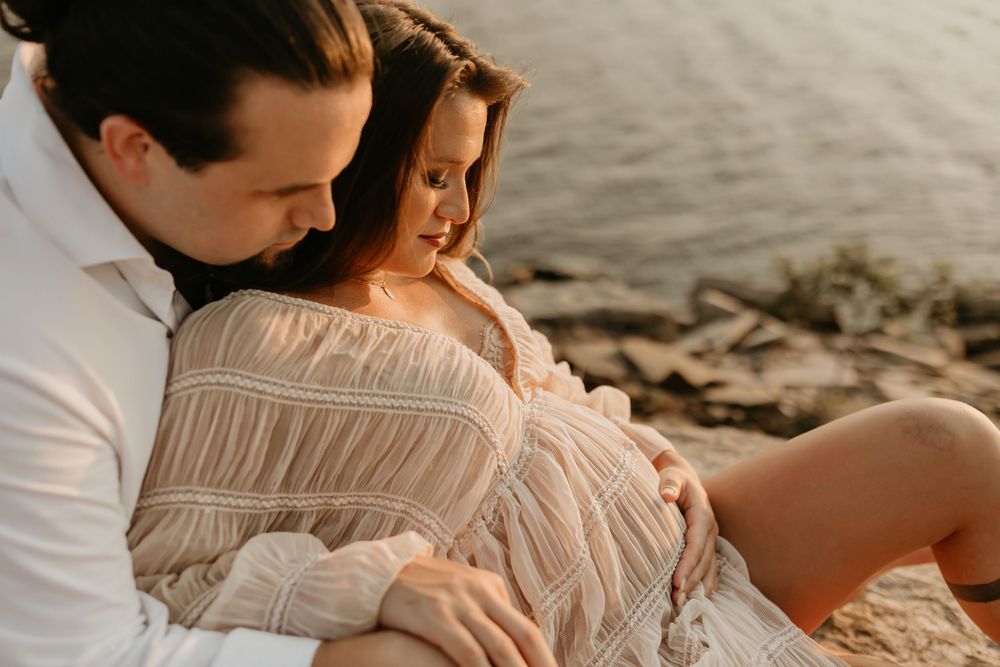 Two individuals embrace tenderly on a beach at sunset, wearing neutral-toned clothing while sitting on the sandy shore.