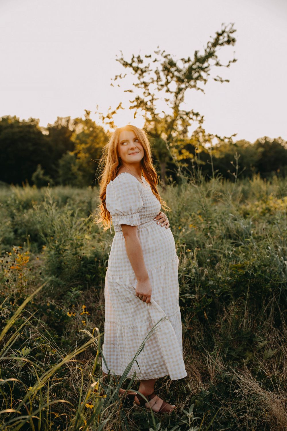 Woman in white gingham dress standing in a golden field at sunset.
