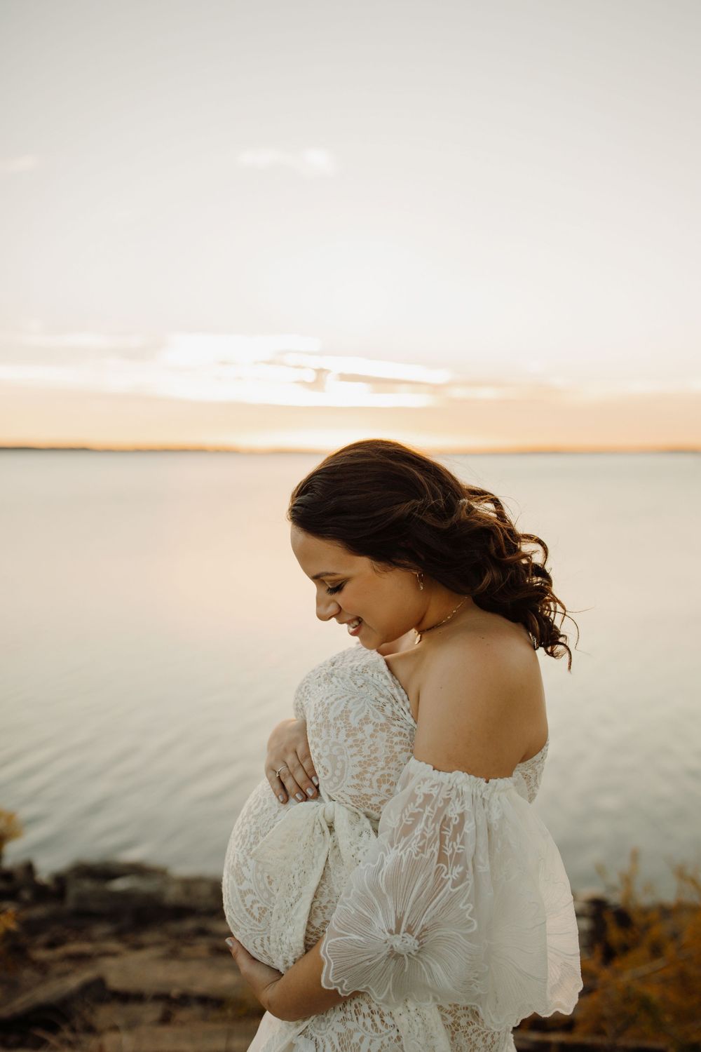 A person in a white lace dress stands by the ocean at sunset with their back turned to the camera.