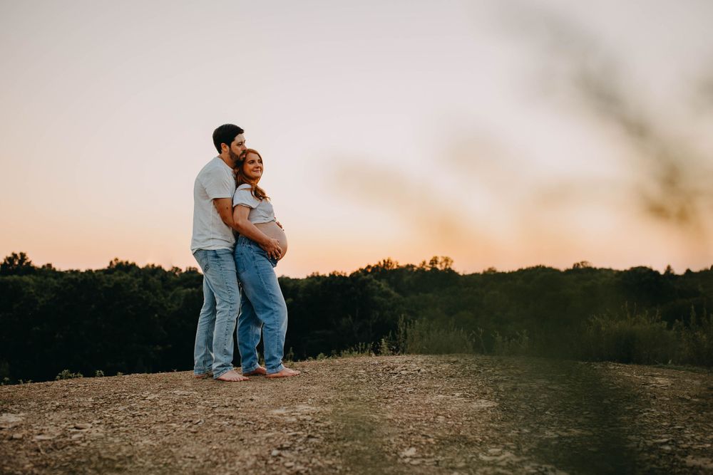 Couple embraces while standing on hilltop against sunset sky in romantic maternity photoshoot.