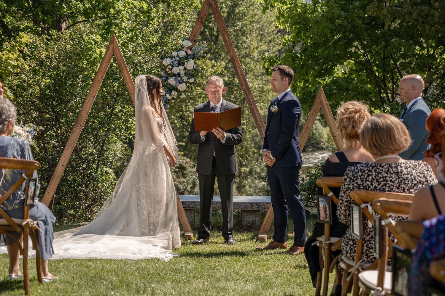 Outdoor wedding ceremony takes place under wooden arch with guests seated on chairs.