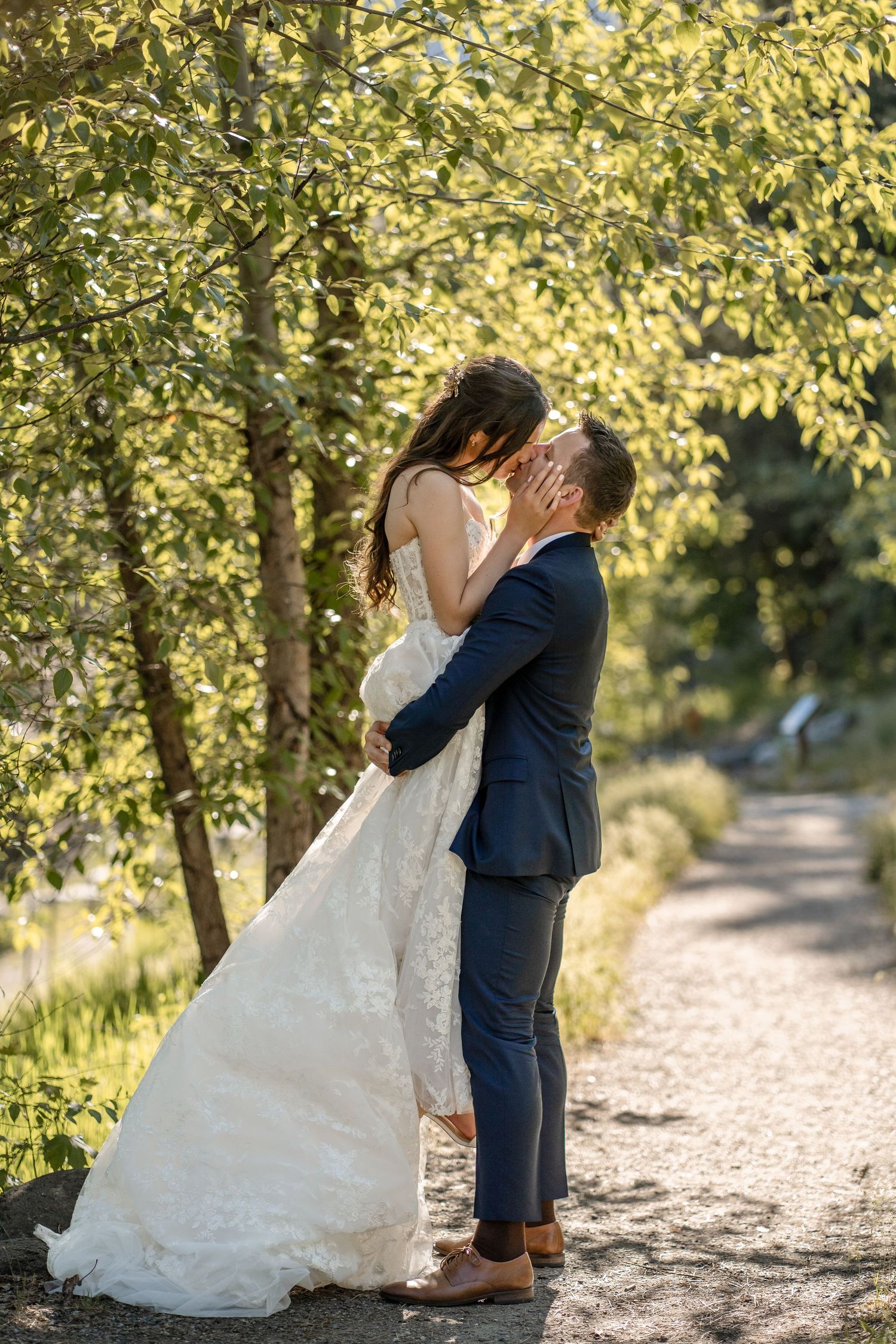 A romantic wedding moment captured on a tree-lined path with natural sunlight filtering through leaves.