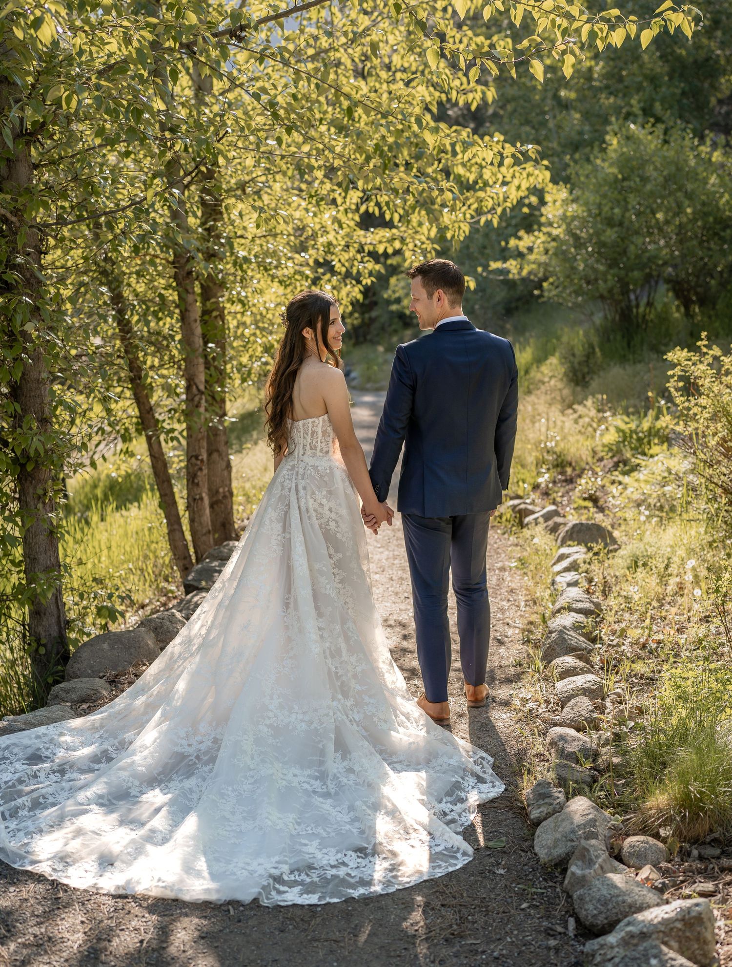 A lace wedding dress train trails beautifully on a nature path as couple walks hand in hand.