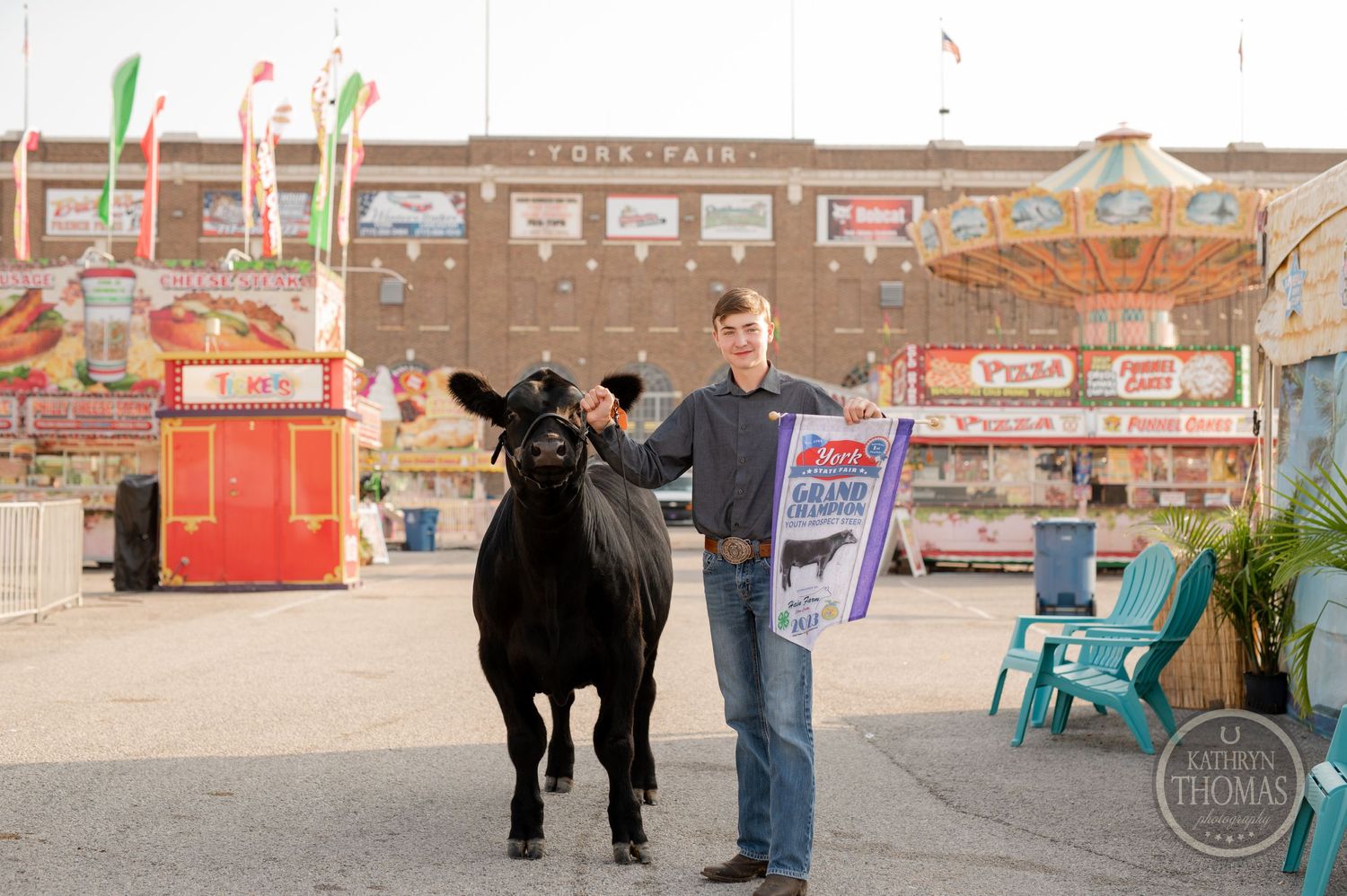 A black show cow and handler pose at a carnival fairground with colorful flags and concession stands.
