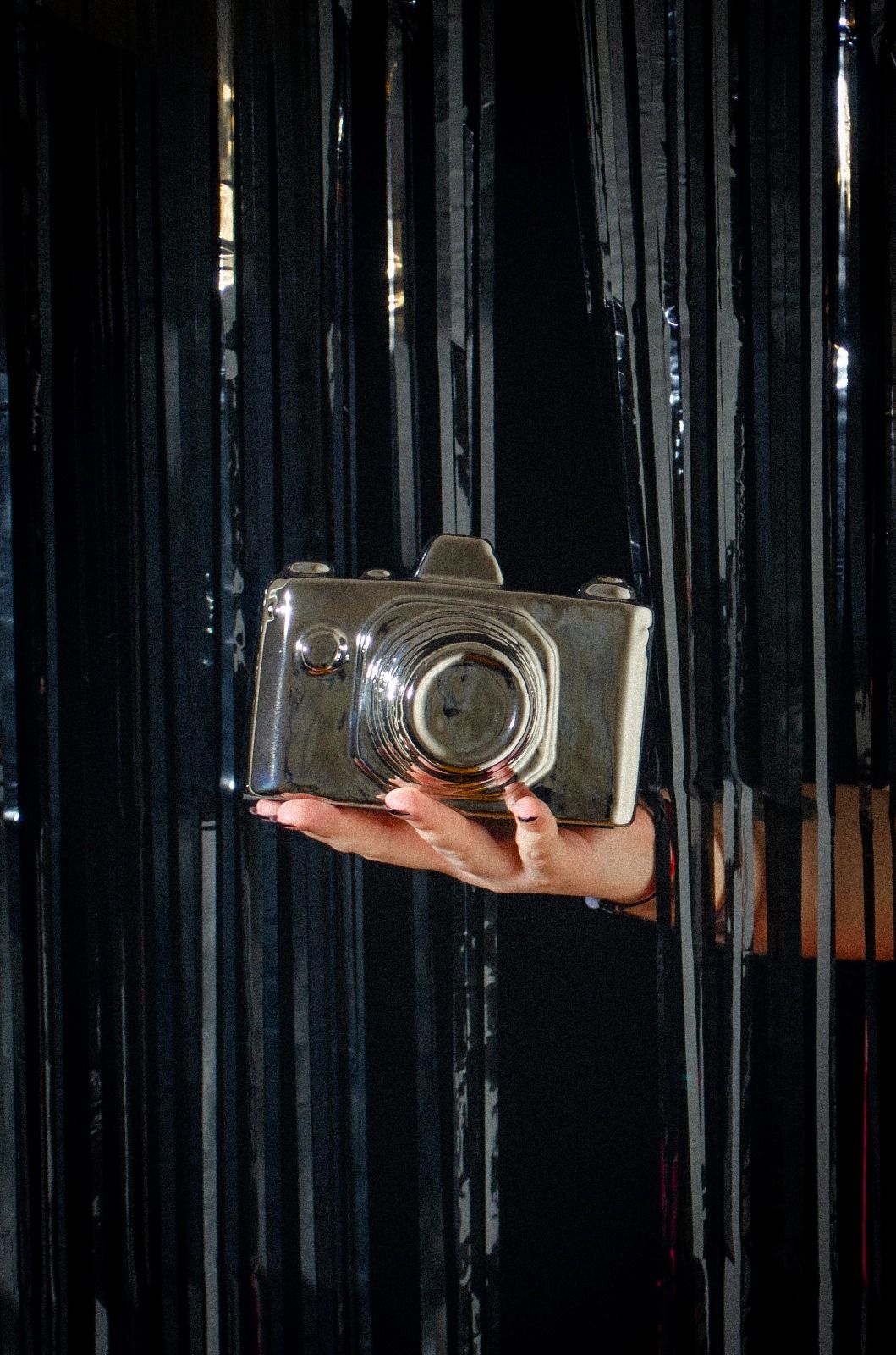 A hand holds a silver camera sculpture against a dark fringe background.