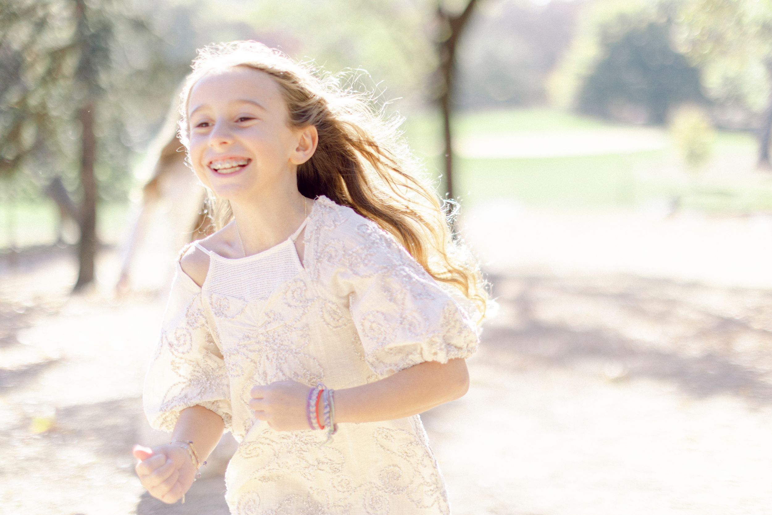 Young person in a white lace dress runs joyfully through a sun-dappled outdoor setting.