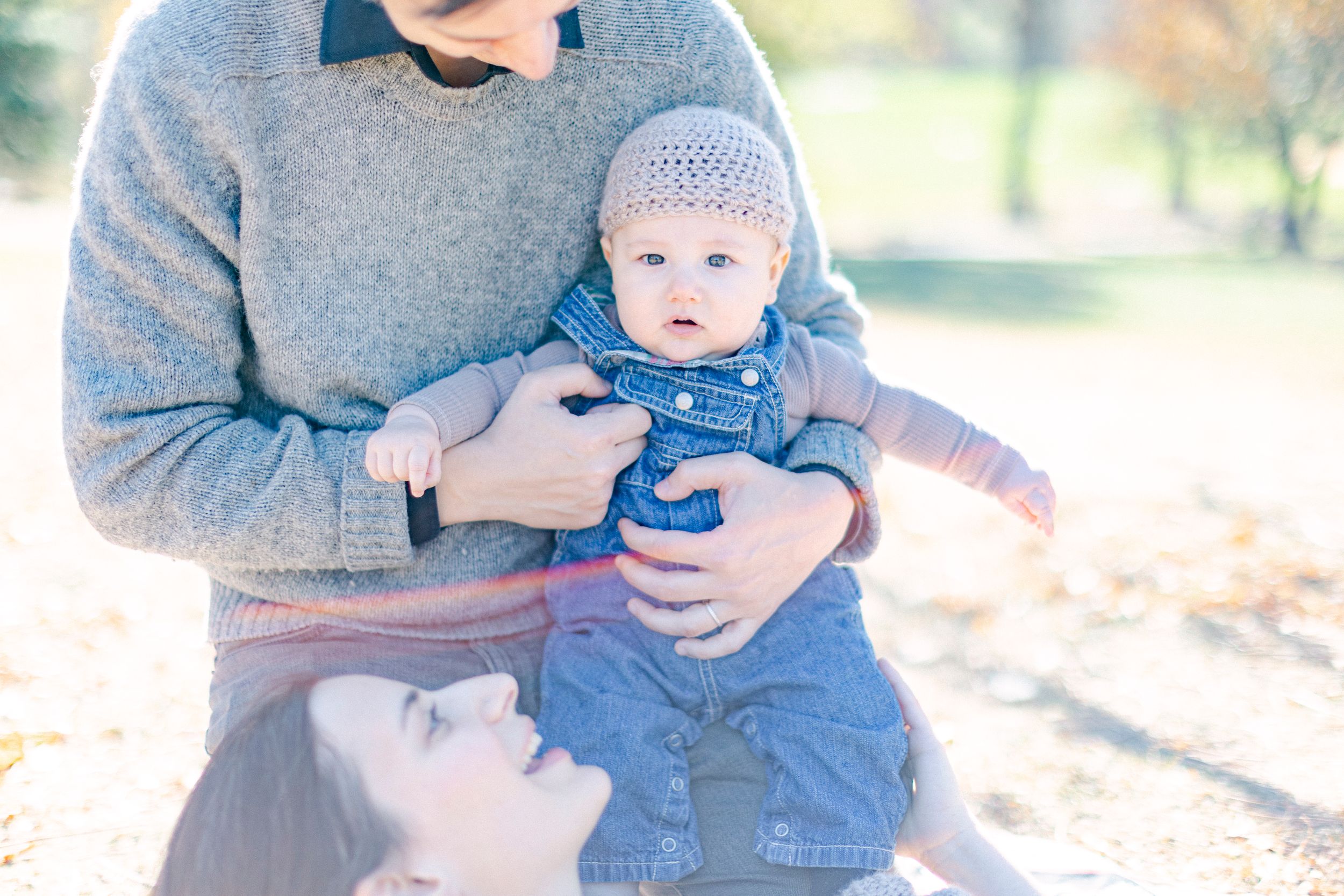A baby in blue overalls and knit cap being held during an outdoor photoshoot.