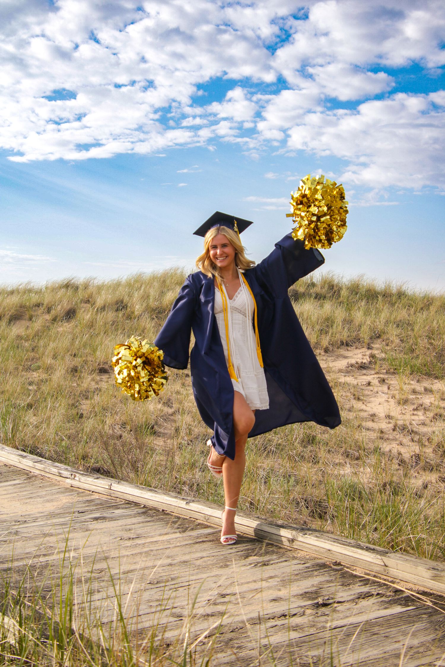 Graduate in navy cap and gown poses playfully with gold pom poms on wooden boardwalk.