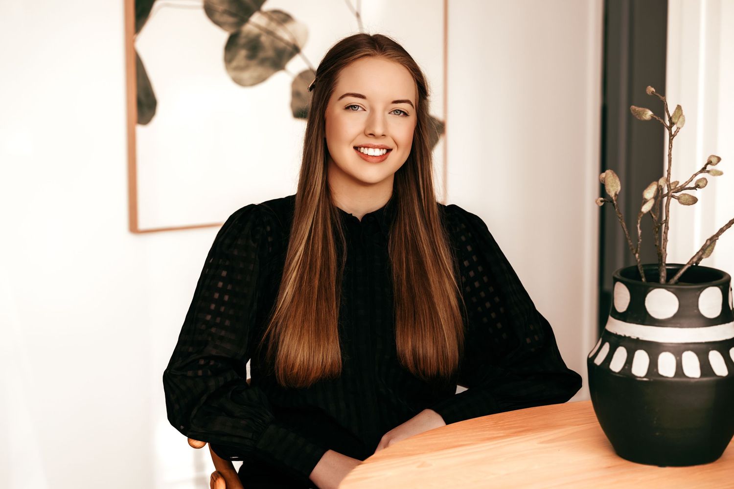 Professional portrait of young person in black top sitting at desk with decor in background.