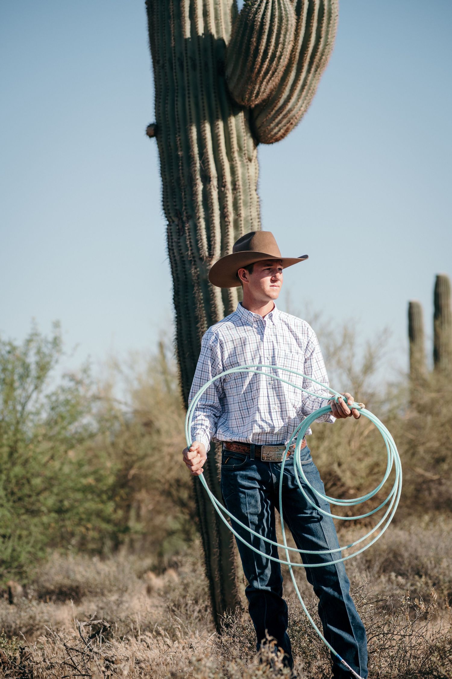 Person in western wear holding lasso poses near majestic saguaro cactus in desert setting.
