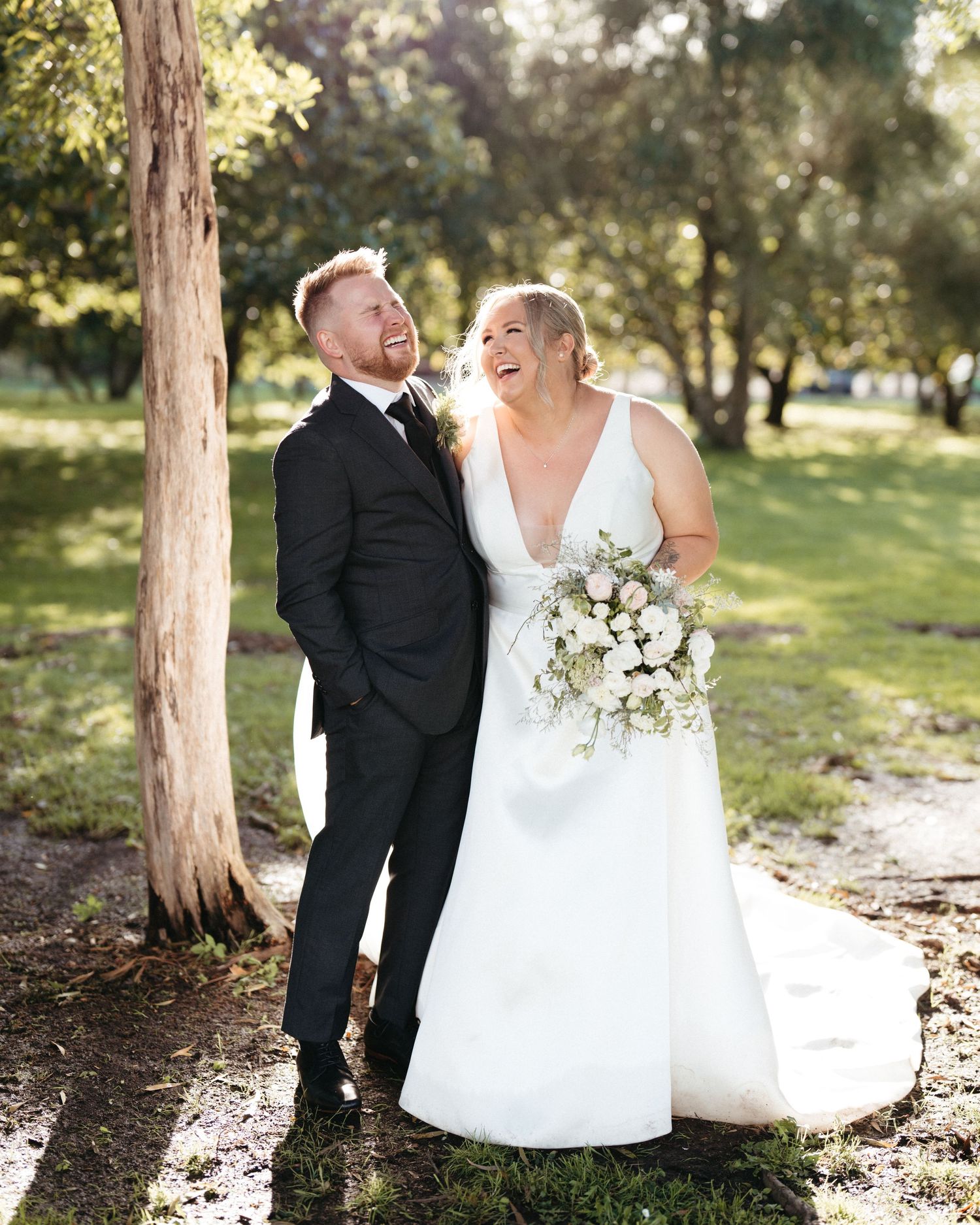 A happy couple shares a joyful moment under a tree in their wedding attire, with a white bouquet in hand at Markovina Vineyard Estate