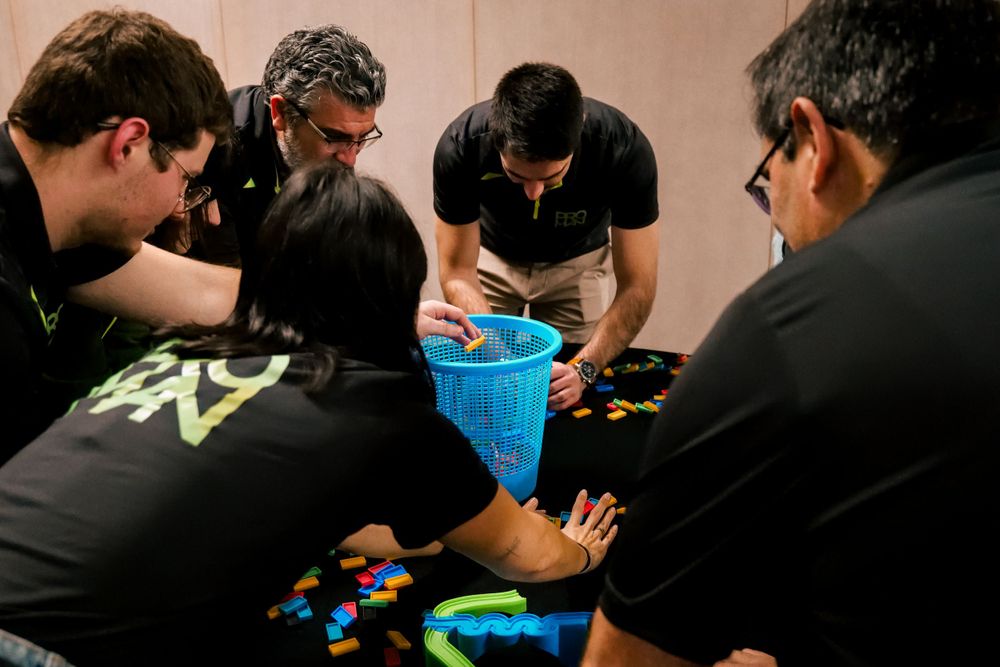 Team members in black shirts huddle around a blue basket with colorful building blocks in a group activity.