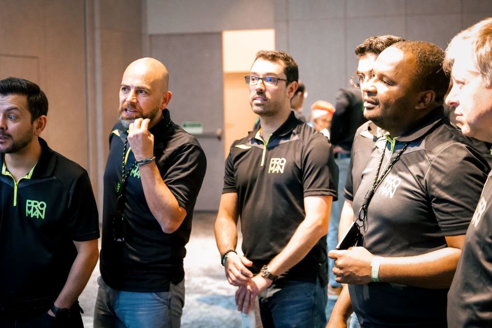 Four men in PROMAN branded black polo shirts attentively listen during a corporate team event.