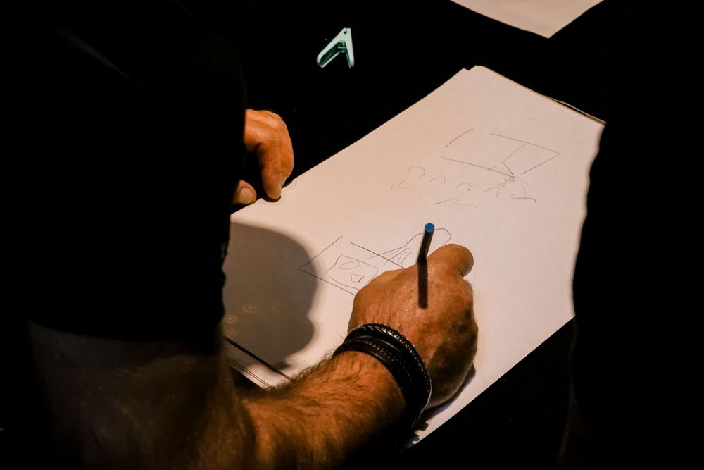 A person in a PROMAN shirt sketches a diagram on white paper with a blue pen in a dark setting.