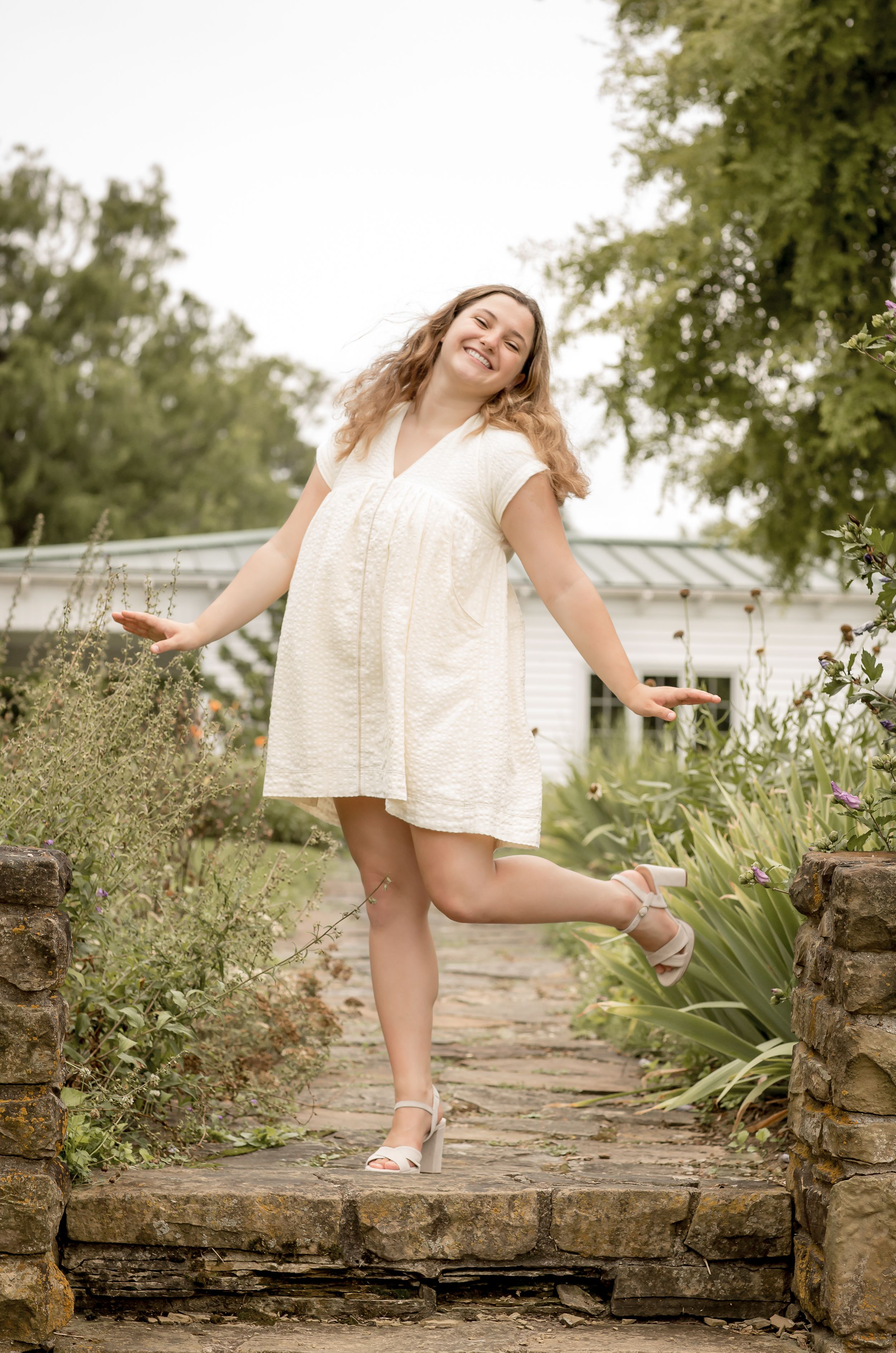 Happy curly-haired girl in a white dress balancing on one foot on a stone path in a garden.