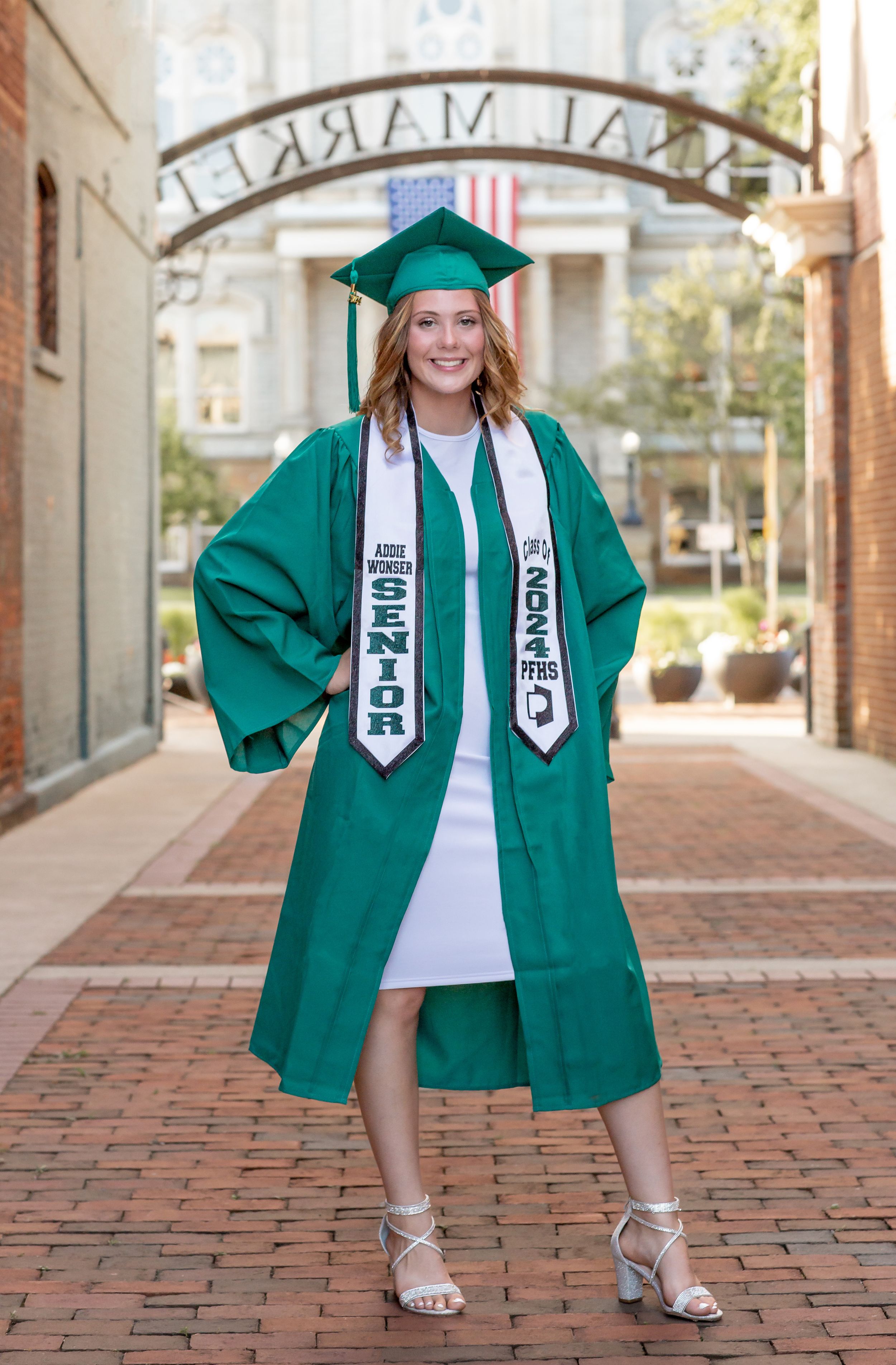 High school senior girl in green graduation cap and gown posing at outdoor market brick walkway.