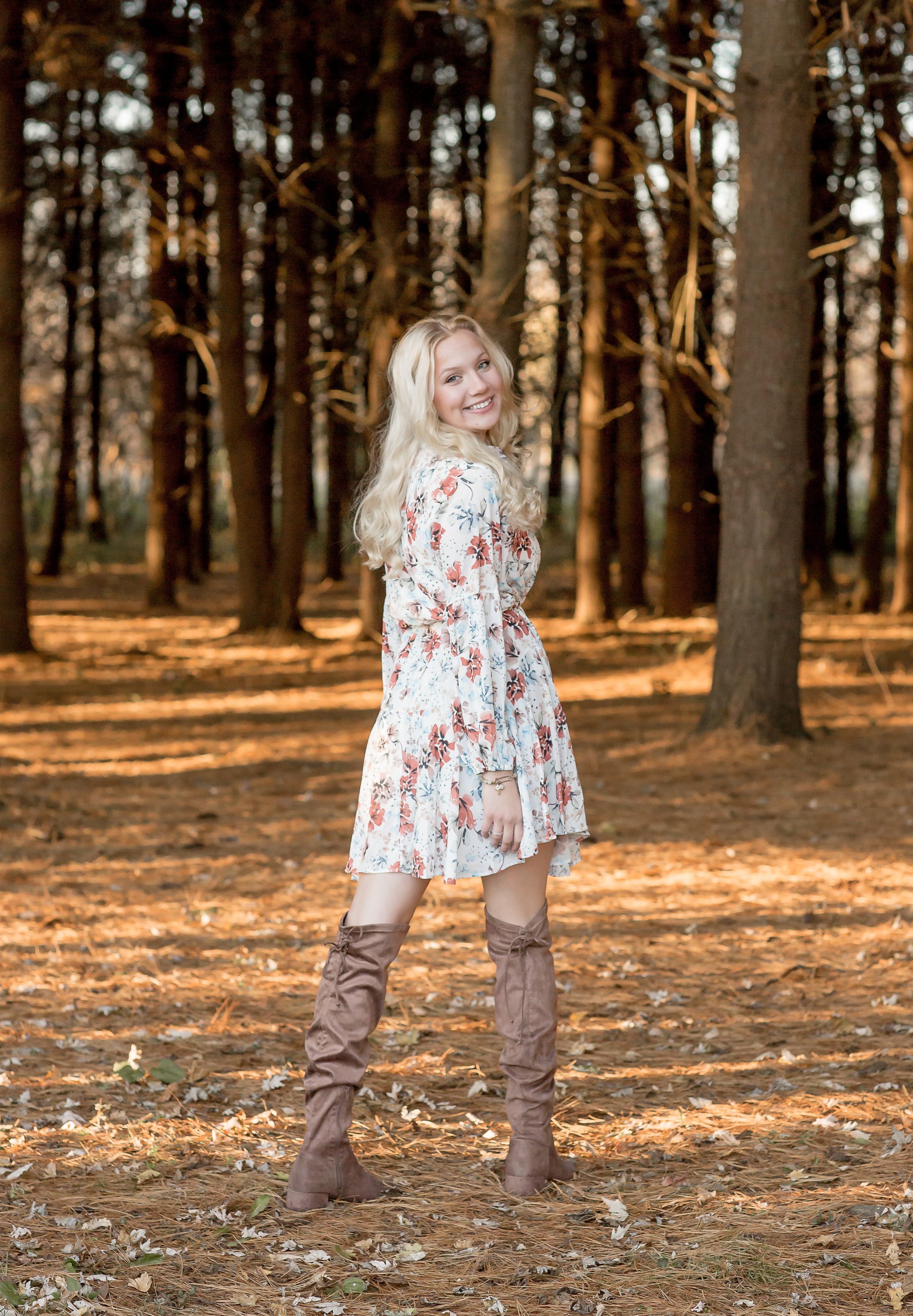 Smiling blonde girl in a floral dress and tall brown boots posing in a sunlit autumn forest.
