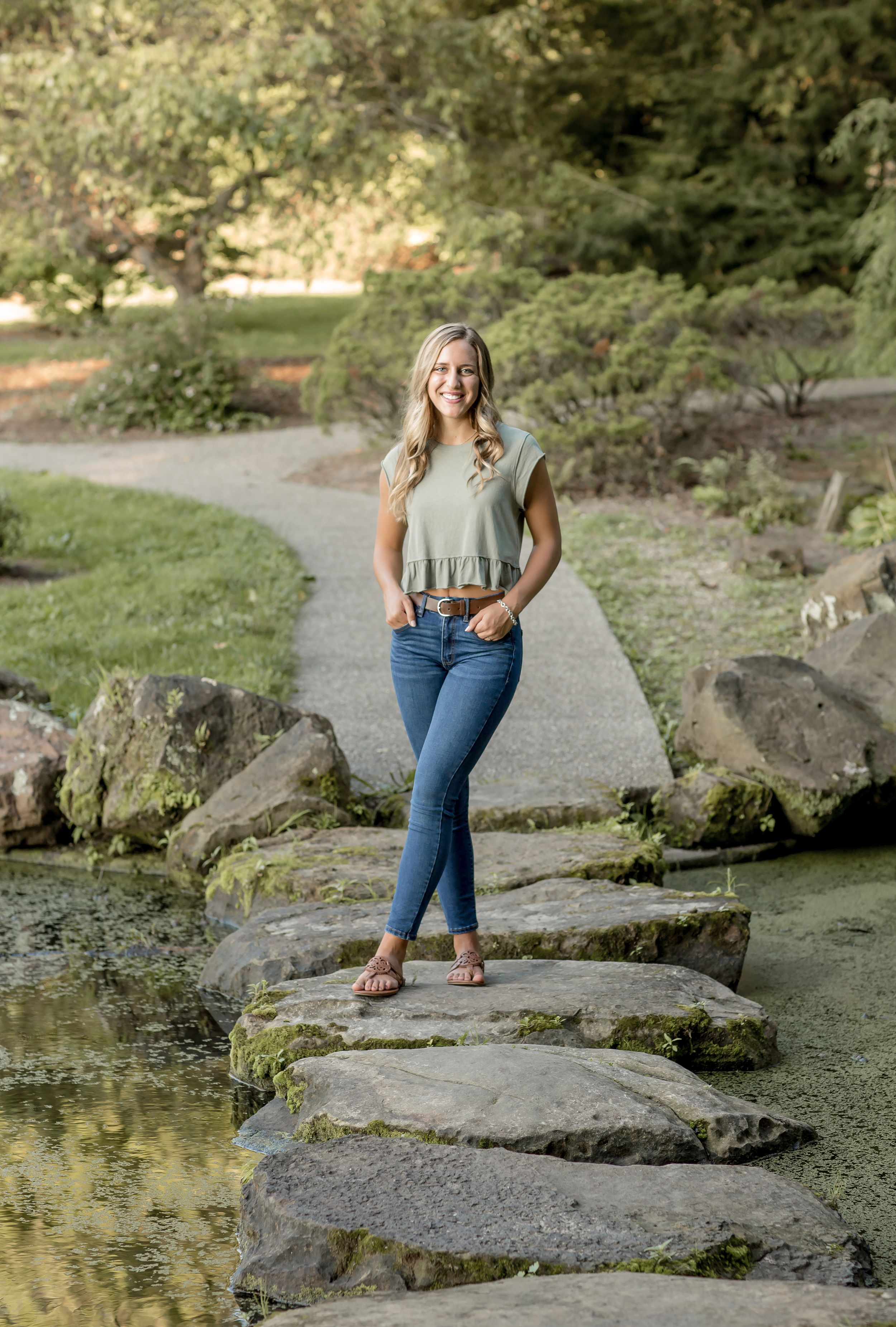 Smiling blonde woman in sage green top and jeans standing on stepping stones.