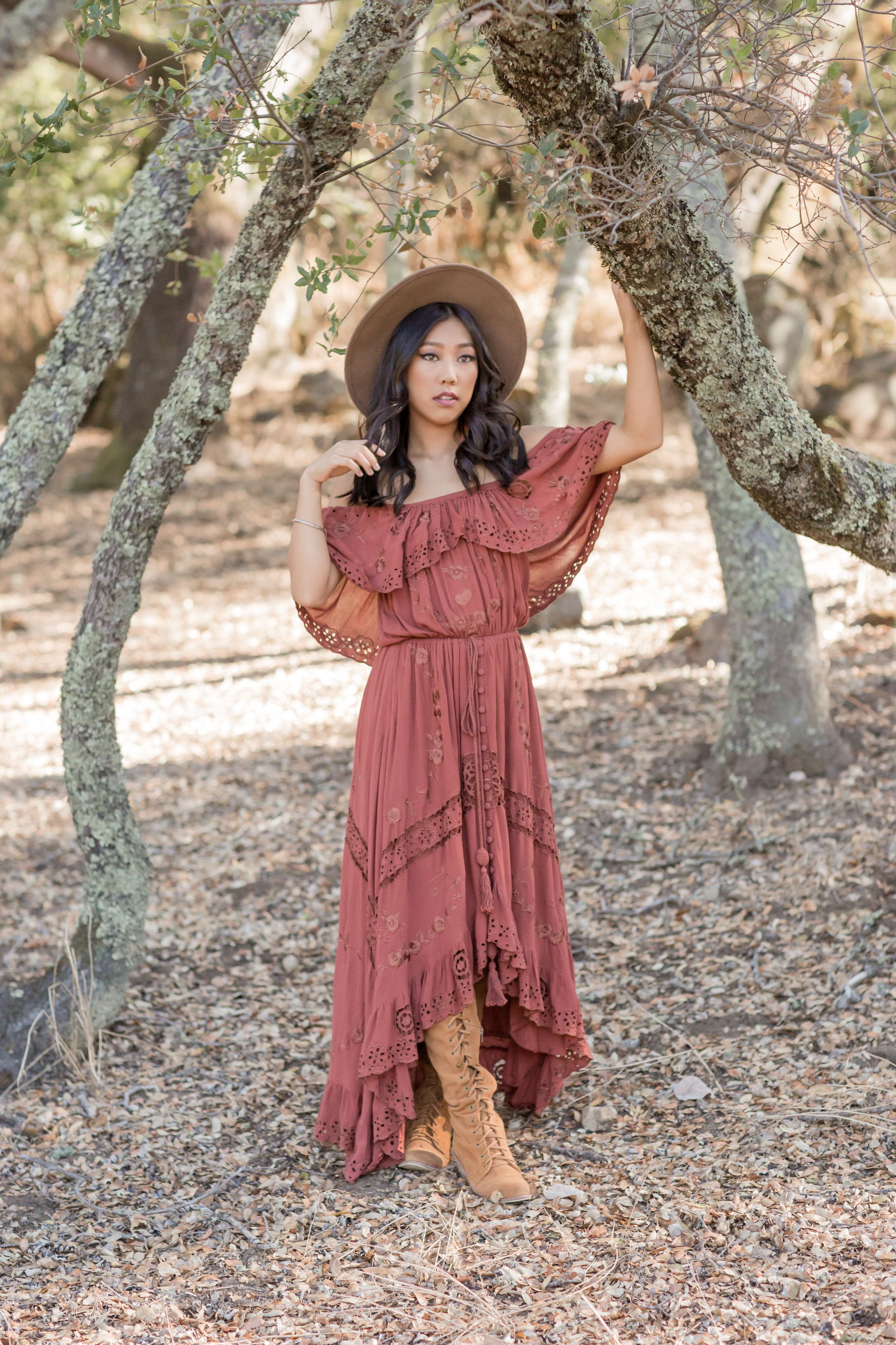 Young woman in boho rust maxi dress and wide-brim hat posing under tree branches outdoors.