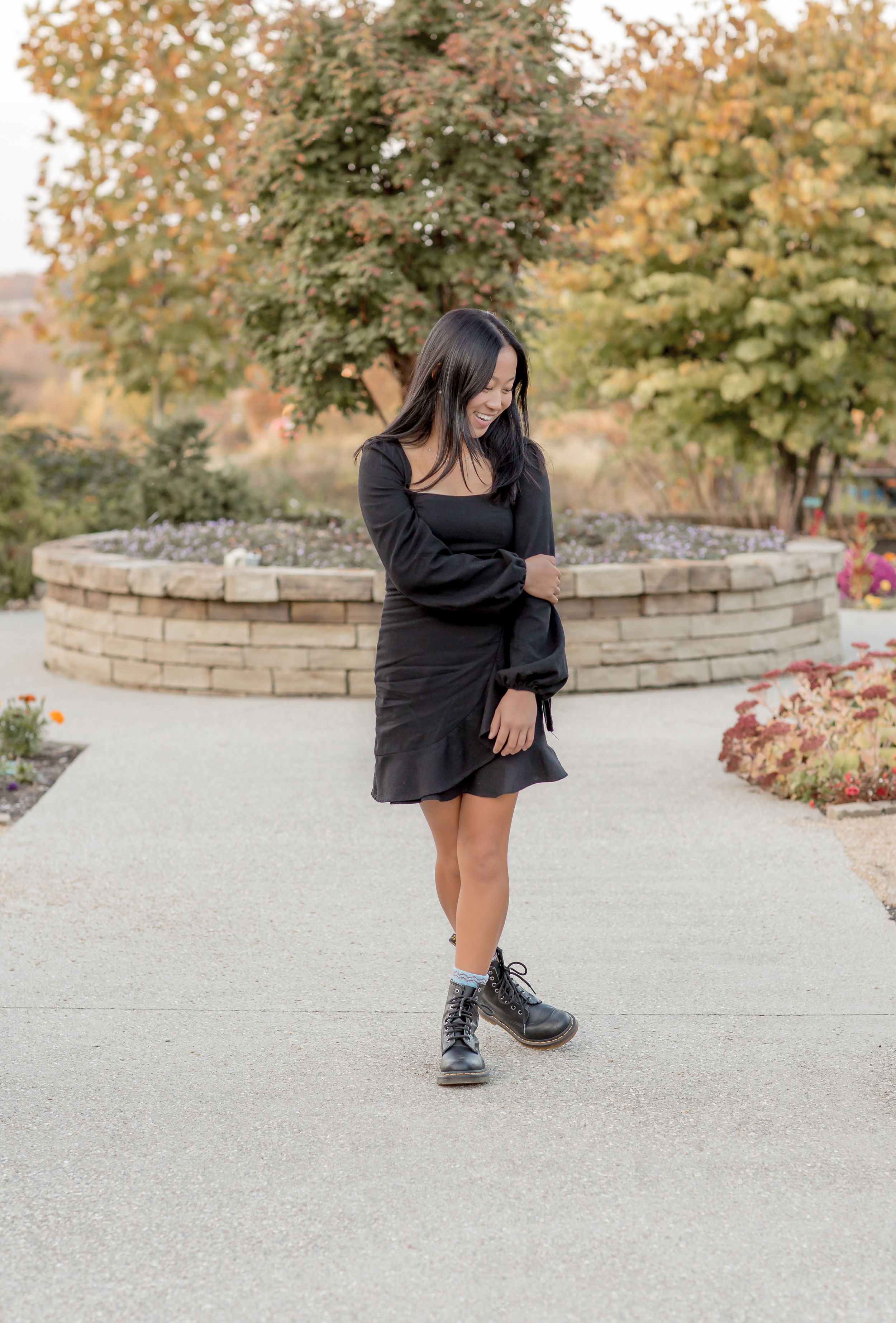 Young woman in a black dress and combat boots looking down while standing on a garden pathway.