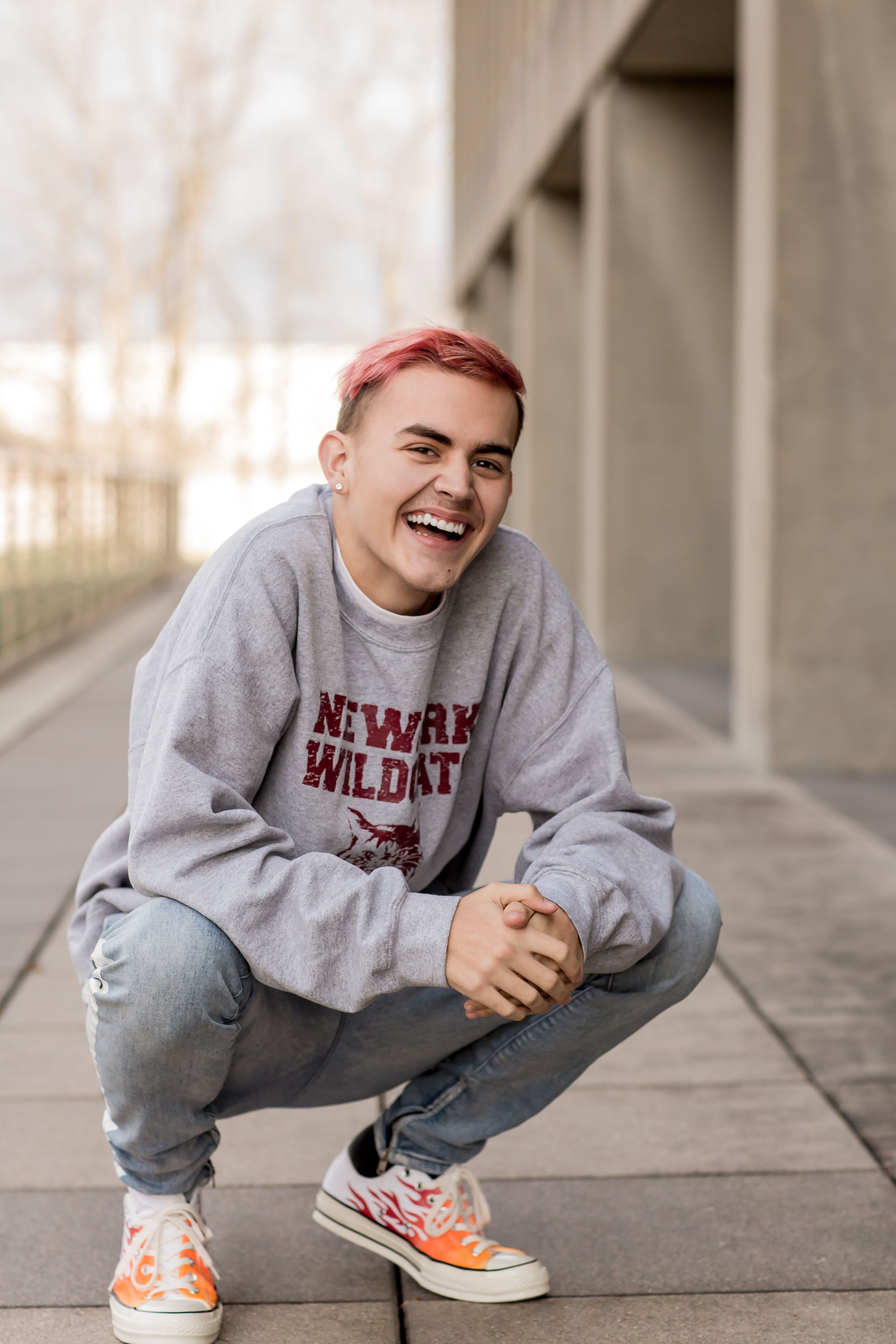 Laughing teen with pink hair in Newark Wildcats sweatshirt crouching on outdoor walkway.