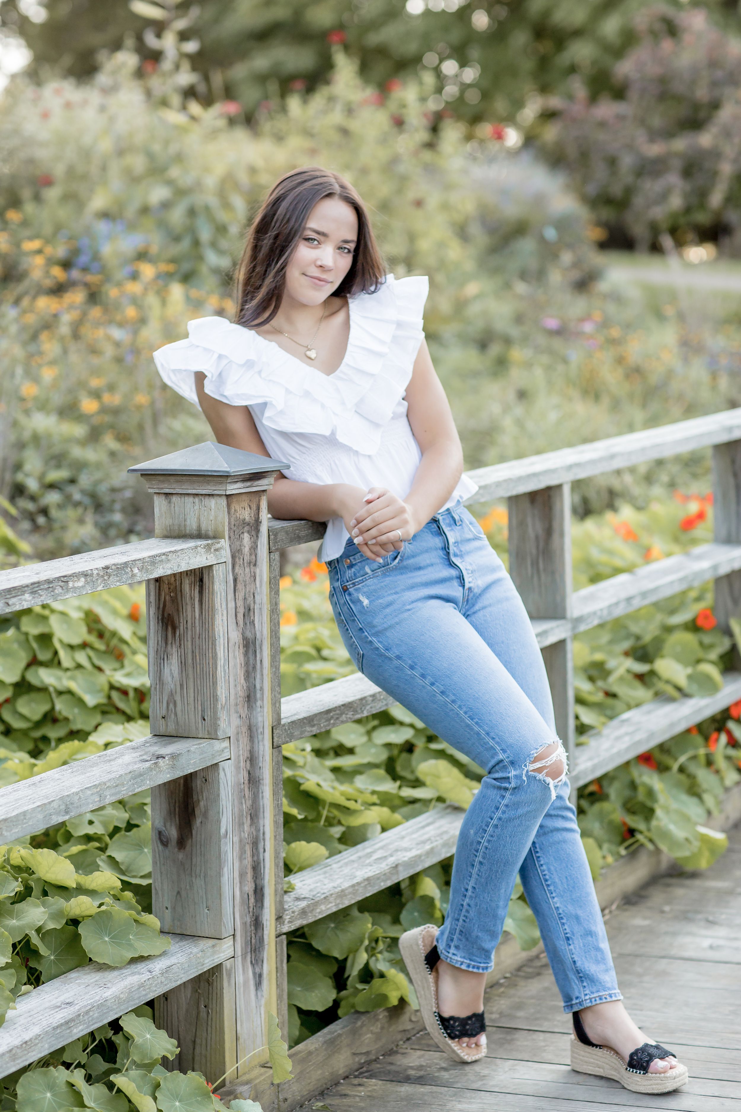 Young brunette woman in white ruffle top and jeans leaning on wooden fence.
