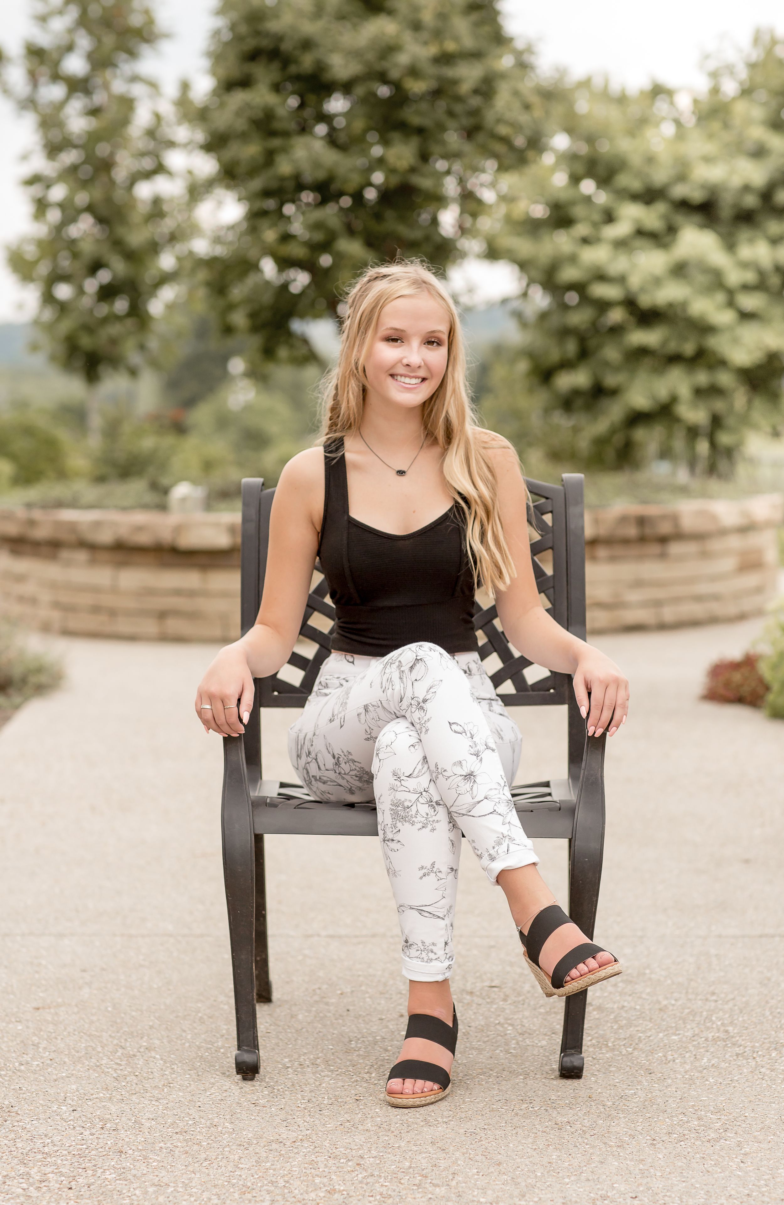 Smiling blonde teen girl sitting on a park bench wearing a black tank top, white marble print leggings, and black sandals.