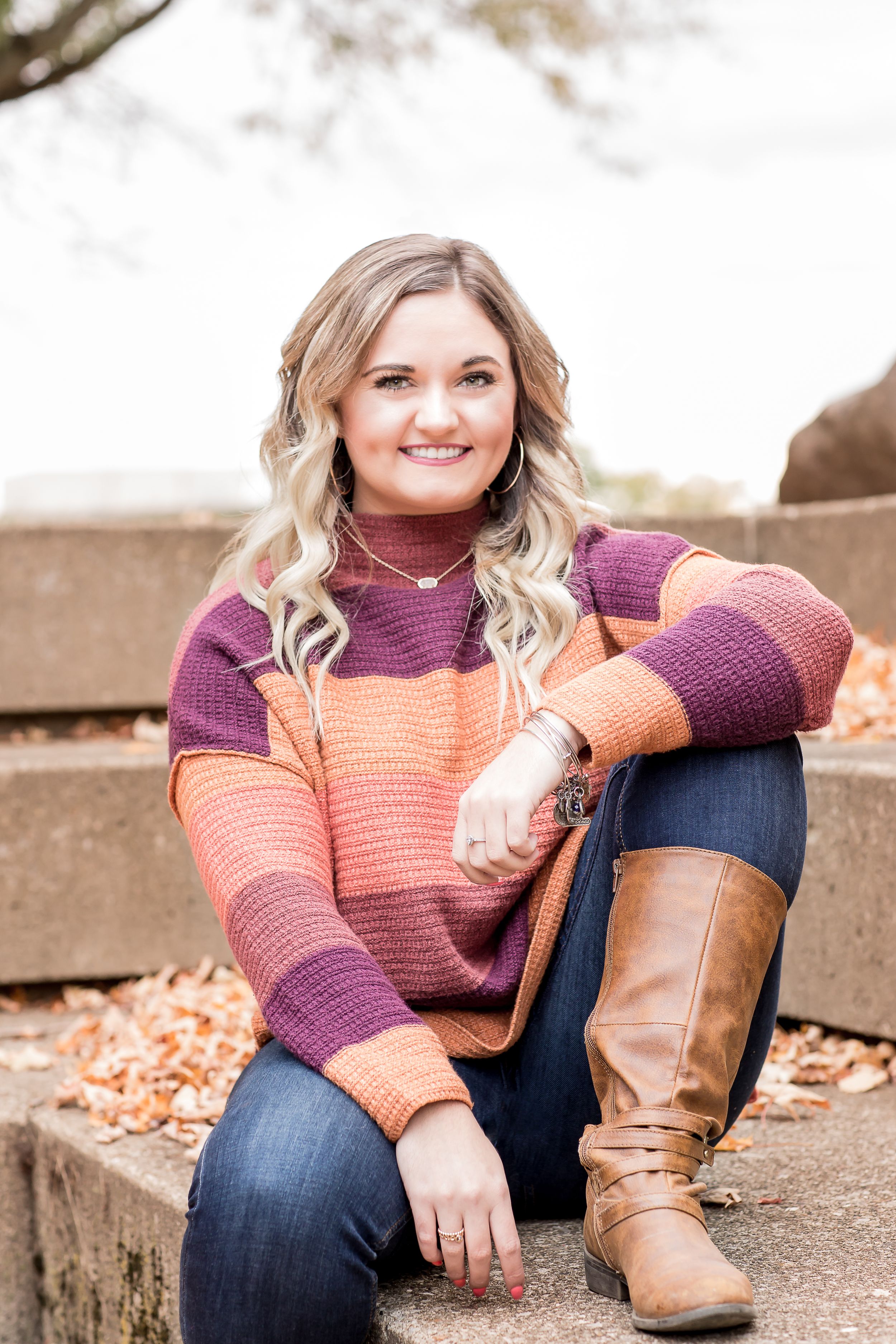 Smiling young woman in colorful striped sweater and boots sitting on stone steps outdoors in fall.