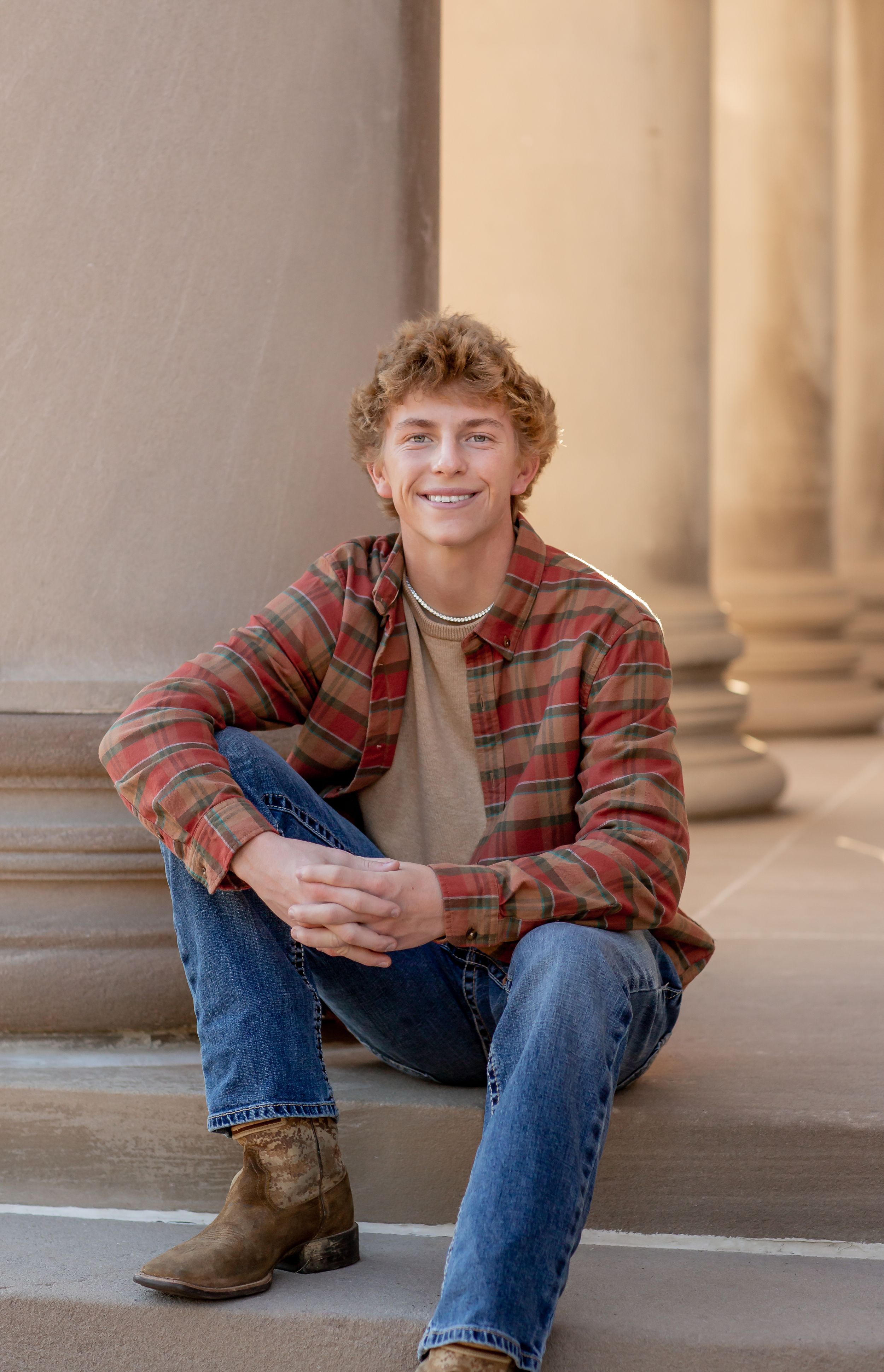 Smiling teen boy in red plaid shirt and jeans sitting on stone steps with columns.