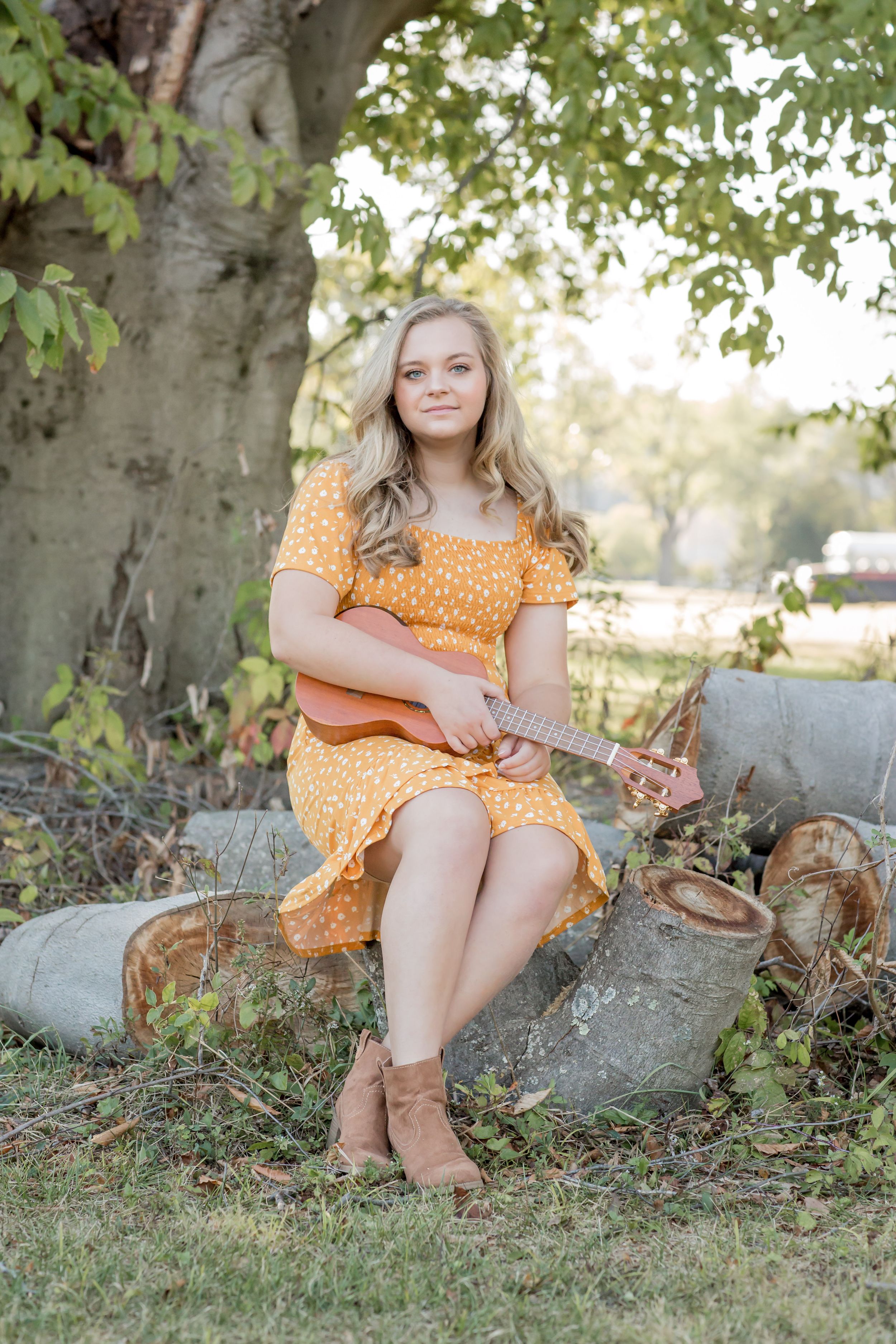 Teenage girl in orange floral dress playing ukulele outdoors under a large tree in natural setting.
