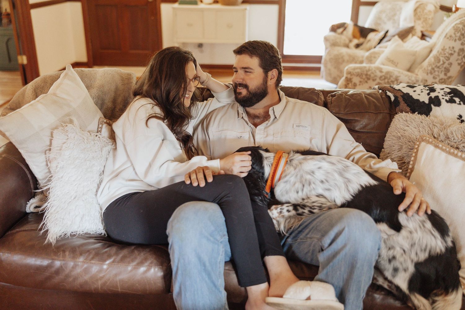Cozy couple lounging on leather couch with throw pillows in home setting.