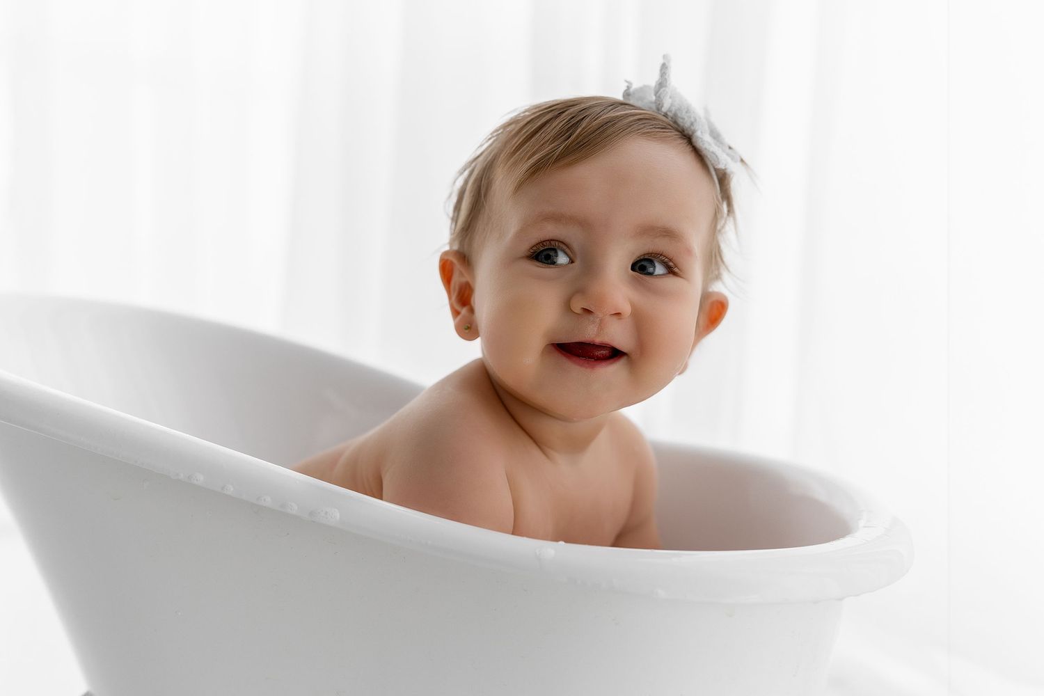 Happy baby taking a bath in white porcelain tub with light curtains behind.