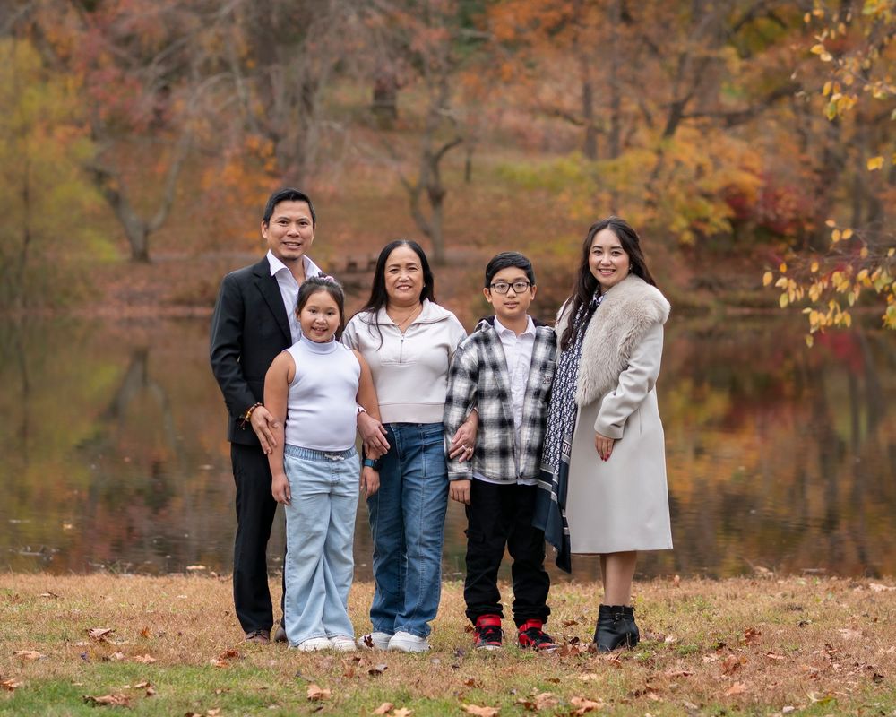 Family of five smiles together outdoors near a pond with vibrant fall foliage backdrop.Holmdel Park NJ
