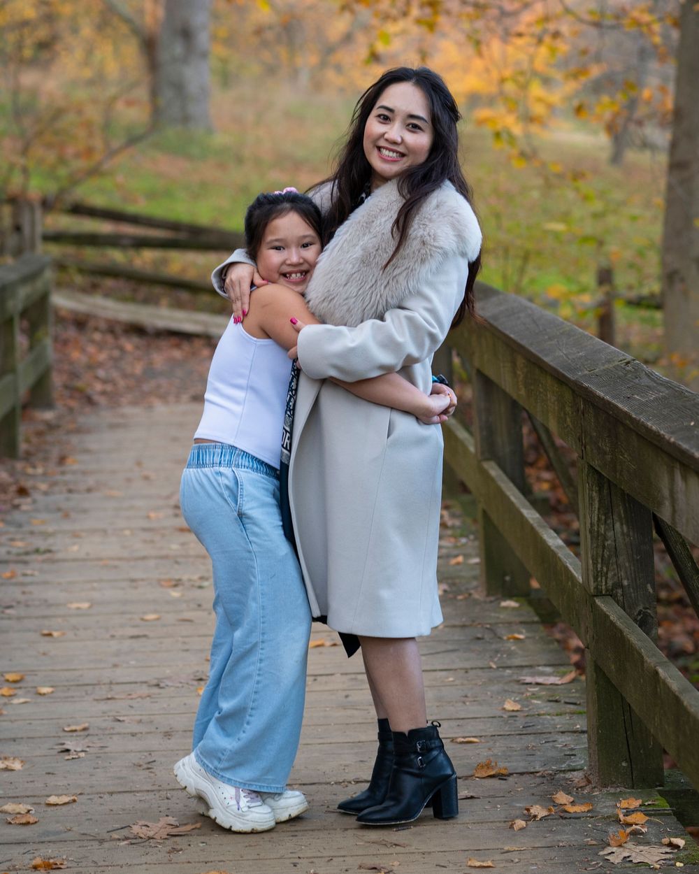 Young girl hugs a smiling woman on a wooden bridge covered in autumn leaves.Holmdel Park NJ
