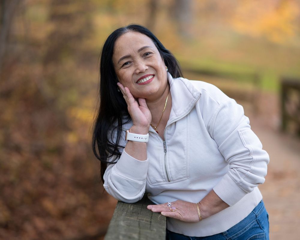 A smiling woman with long dark hair poses outdoors in a white jacket among autumn foliage.Holmdel Park NJ