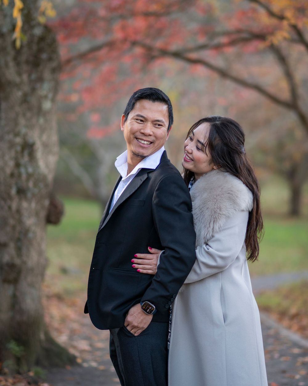 A smiling couple embraces outdoors with vibrant red autumn foliage in the background.Holmdel Park NJ