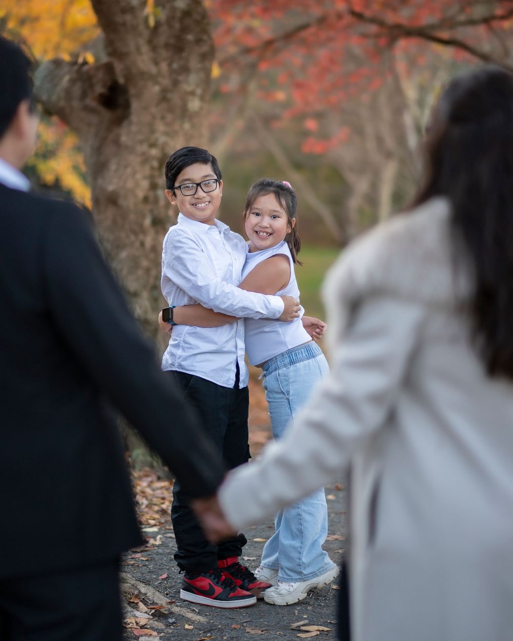 Two children hug and smile on a fall park path while adults hold hands nearby.Holmdel Park NJ