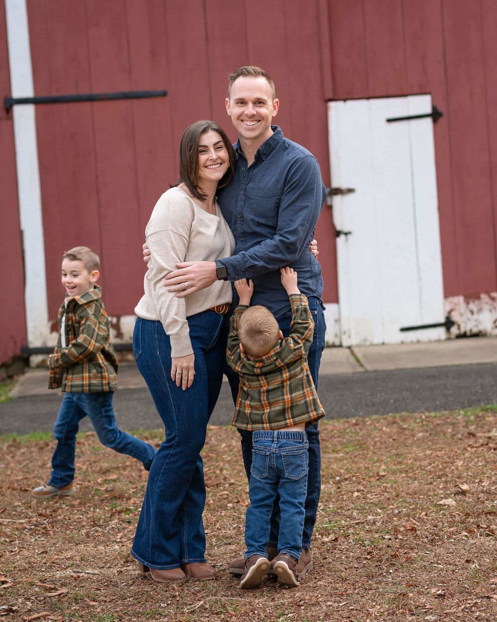Happy family of four poses and laughs together outside a red barn in autumn.Middletown NJ