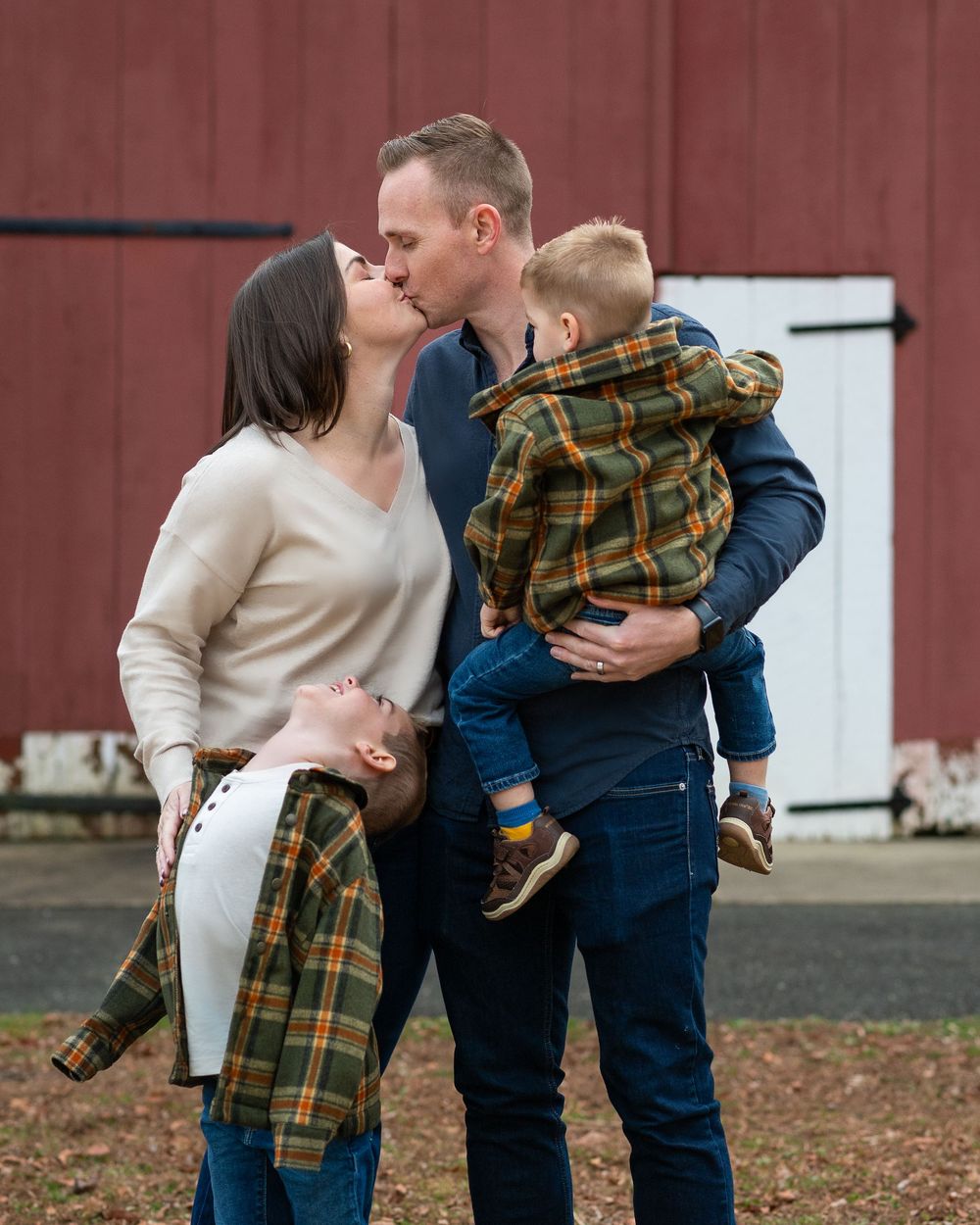Couple shares a kiss while holding their young children outside a red barn. Middletown NJ