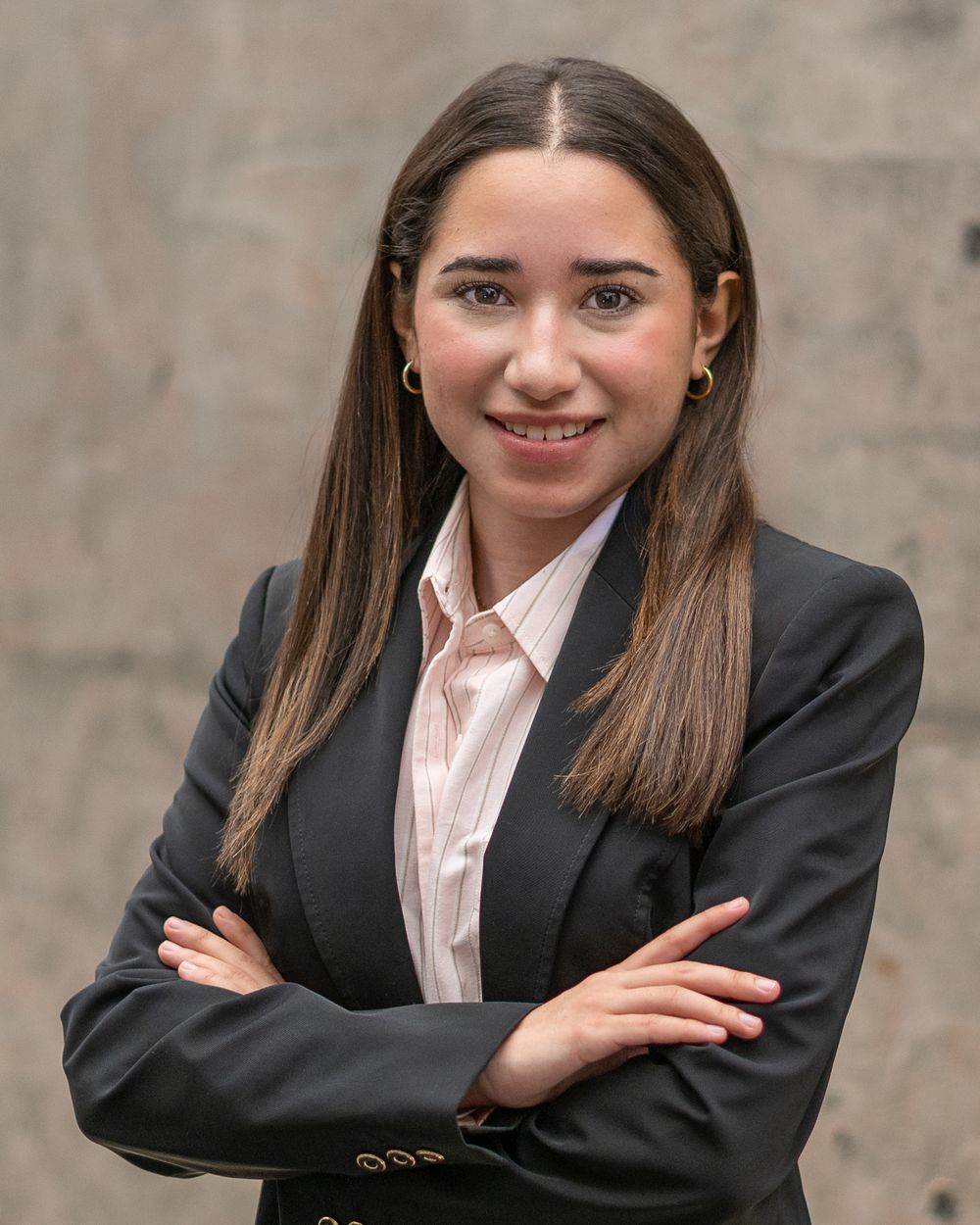headshot of young professional woman in black blazer with arms crossed smiling against a stone wall background. Holmdel NJ