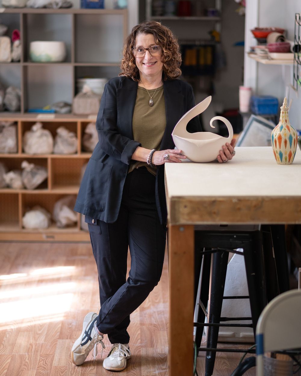 Woman in black blazer holding ceramic sculpture while leaning on table in pottery studio.Red Bank NJ