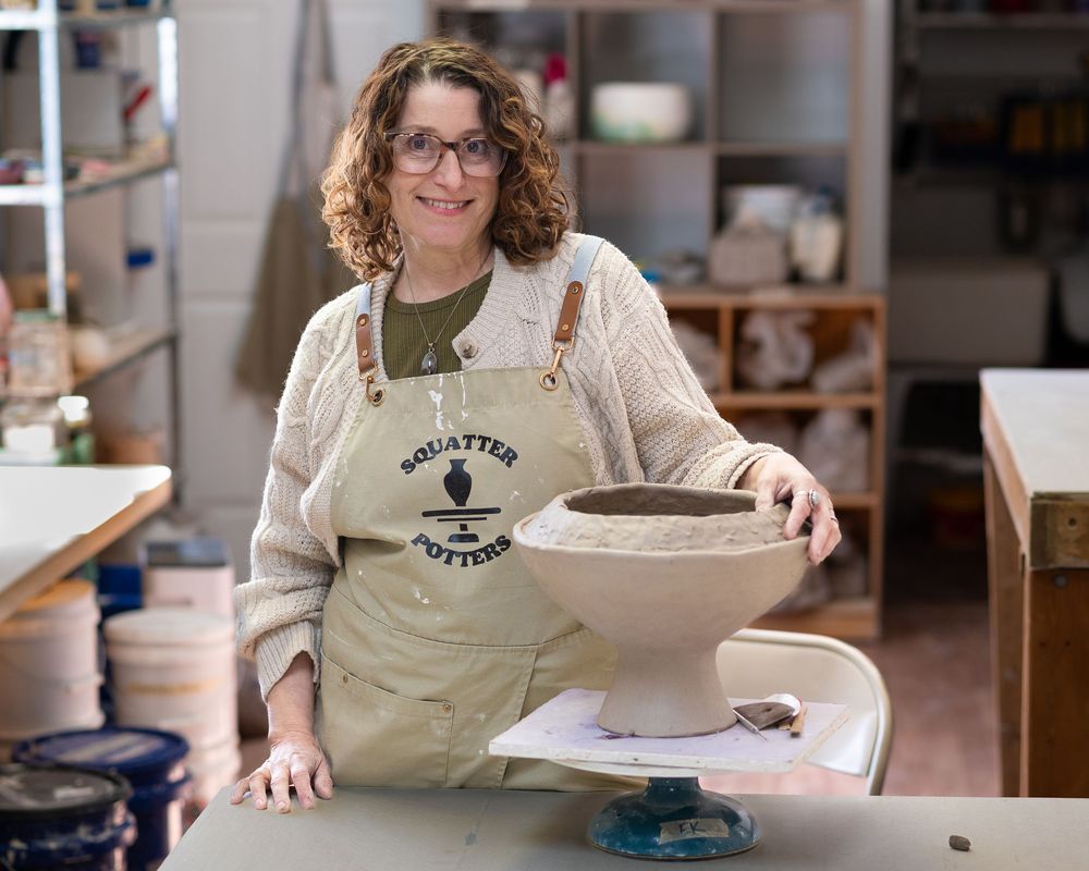Smiling woman in Squatter Potter apron posing with ceramic bowl on wheel in studio.Red Bank NJ