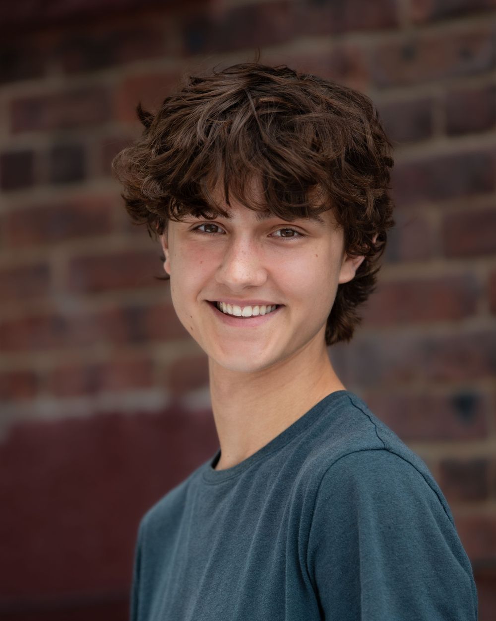 Headshot Portrait of a smiling teen boy in blue shirt against brick background in Atlantic Highlands NJ