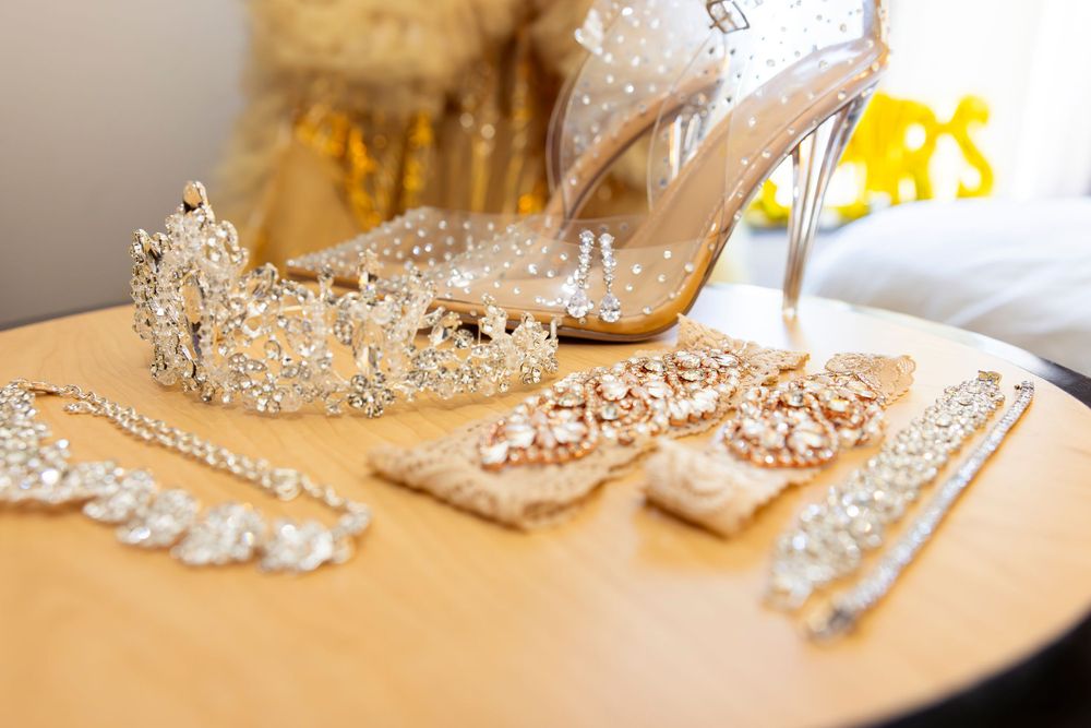 Wedding accessories including crystal tiara, jewelry and silver high heel shoe arranged on beige surface.