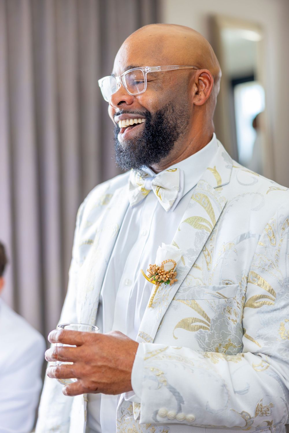 A joyful person wearing a white and gold patterned suit jacket with clear glasses and a bow tie.