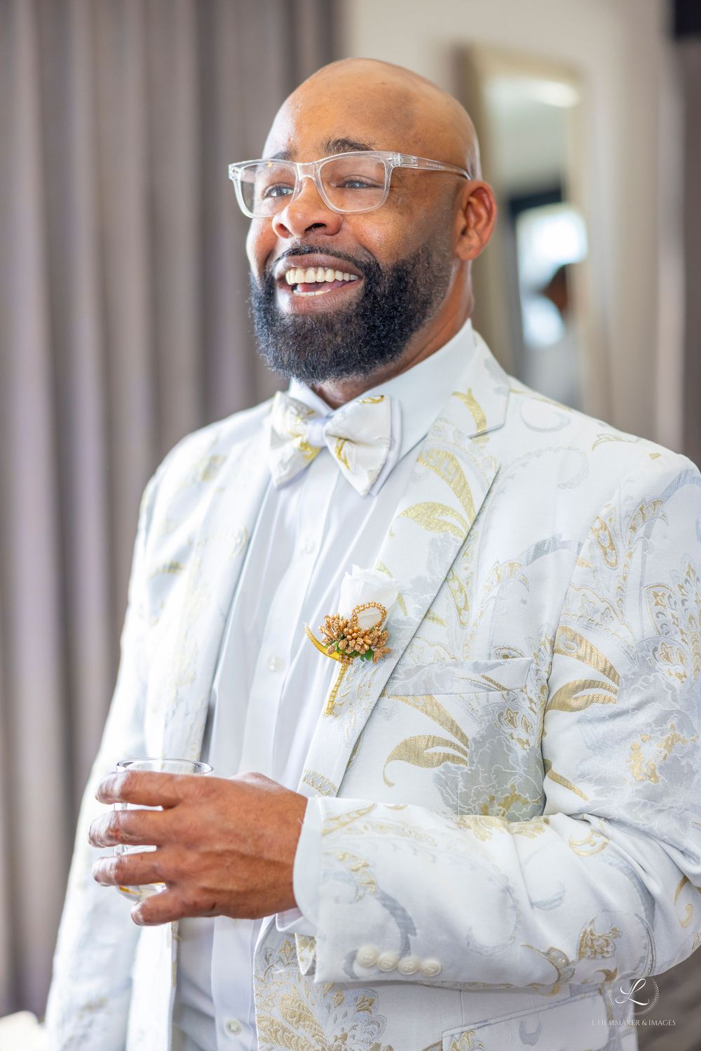 A person in a white and gold patterned suit smiling warmly while wearing clear glasses.