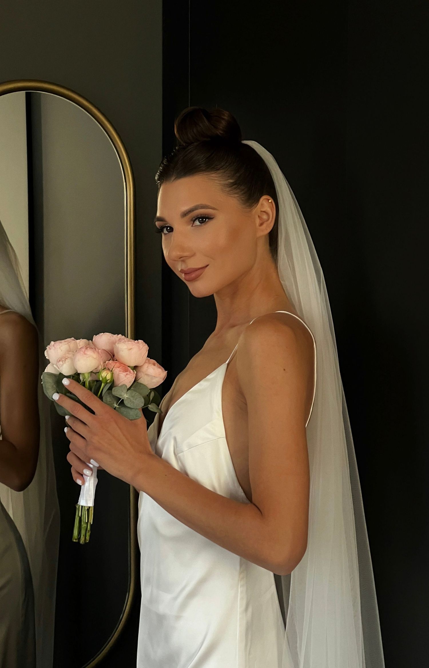 Elegant bride in white dress holding pink roses against dark background with gold accent mirror.