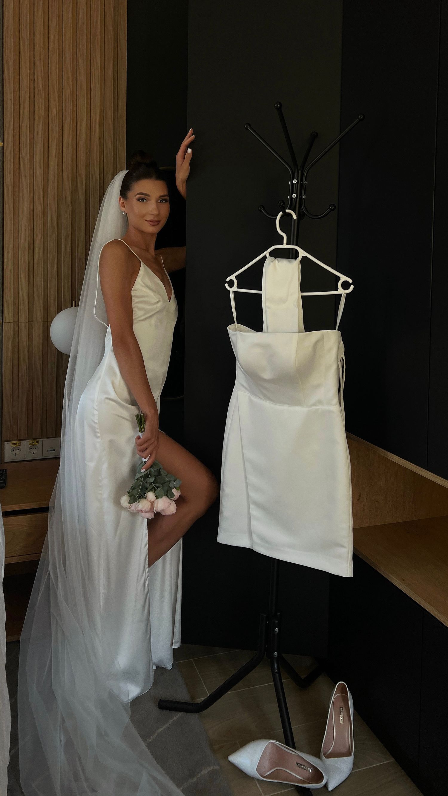 Wedding dress displayed on hanger with bride holding bouquet in background.